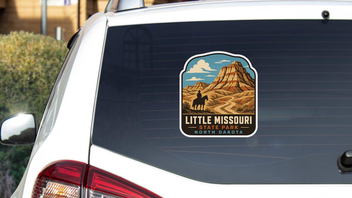 A sticker on the rear window of a white car, depicting a scenic landscape with mountains and a person on horseback, representing Little Missouri State Park in North Dakota.