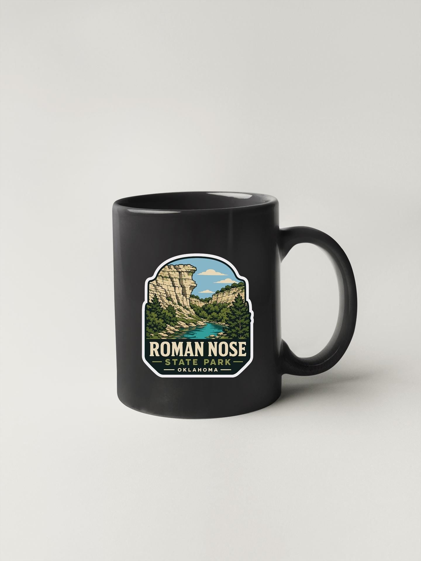 A black coffee mug with a graphic design depicting a scenic landscape, including mountains, a lake, and a waterfall.