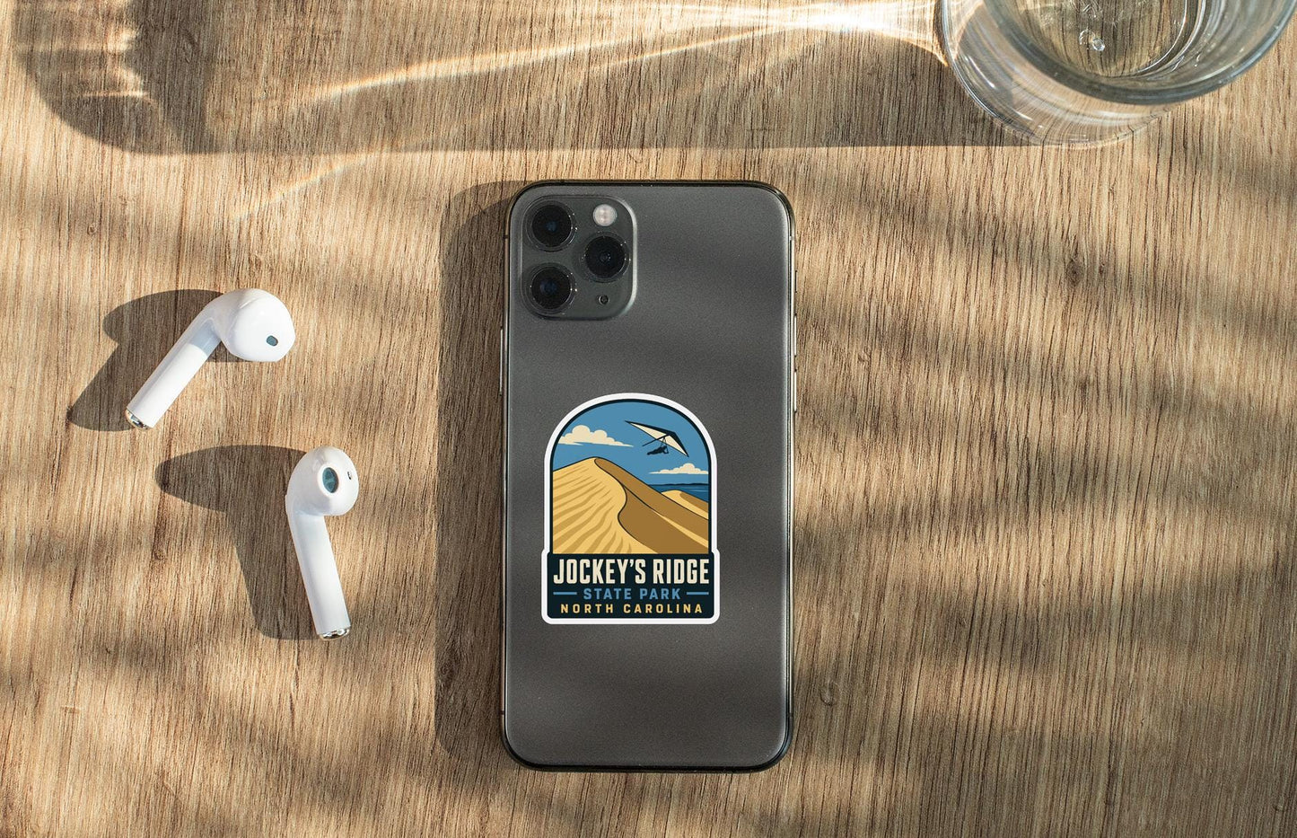 a smartphone with a sticker on the back, placed on a wooden surface alongside a pair of white earbuds and a glass of water.