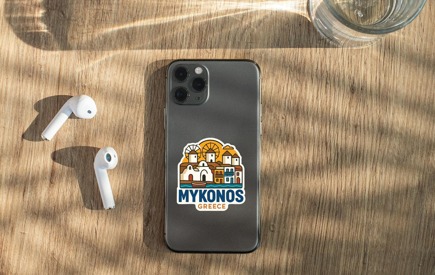 a smartphone with a sticker of a Greek island, placed on a wooden surface alongside a pair of white earbuds and a glass of water.
