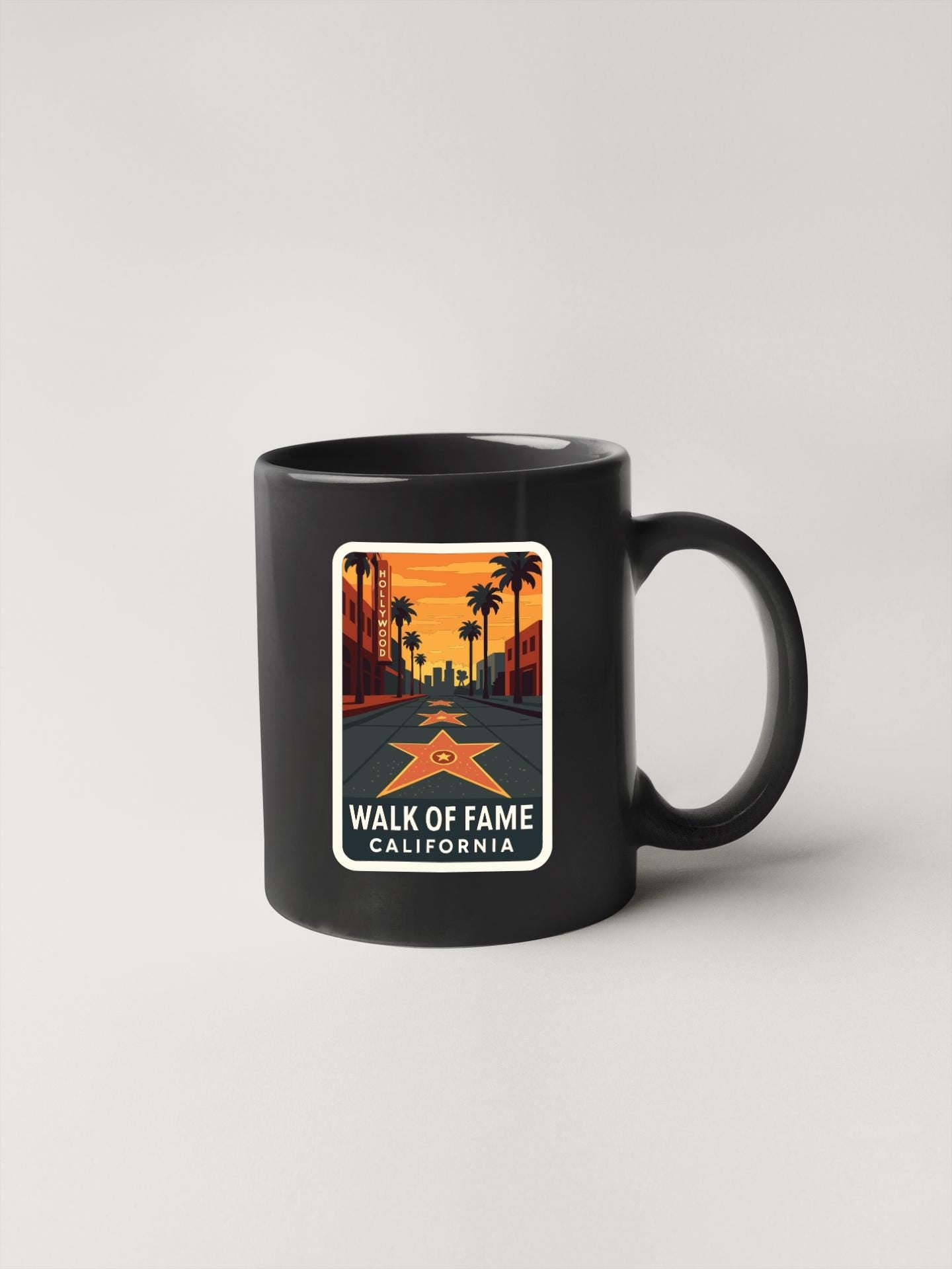 A black coffee mug with a sticker depicting a street scene with palm trees and a star, along with the text "WALK OF FAME CALIFORNIA".