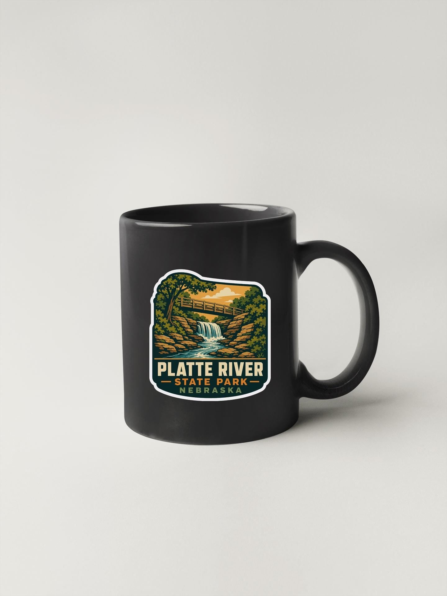 A black coffee mug with a label that says "PLATTE RIVER STATE PARK" and features an illustration of a waterfall and forest.