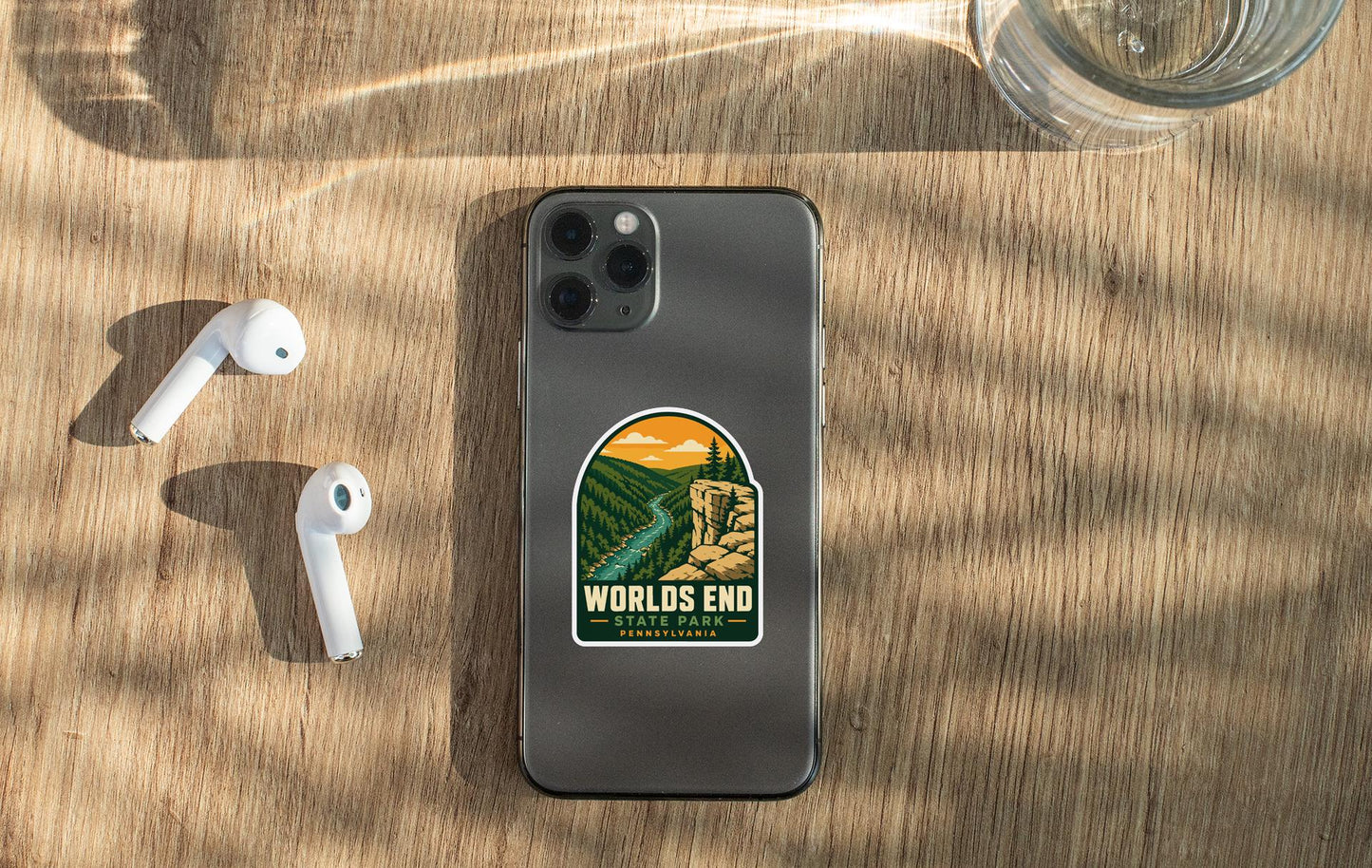 a smartphone with a sticker on the back, along with a pair of white earbuds and a glass of water on a wooden surface.
