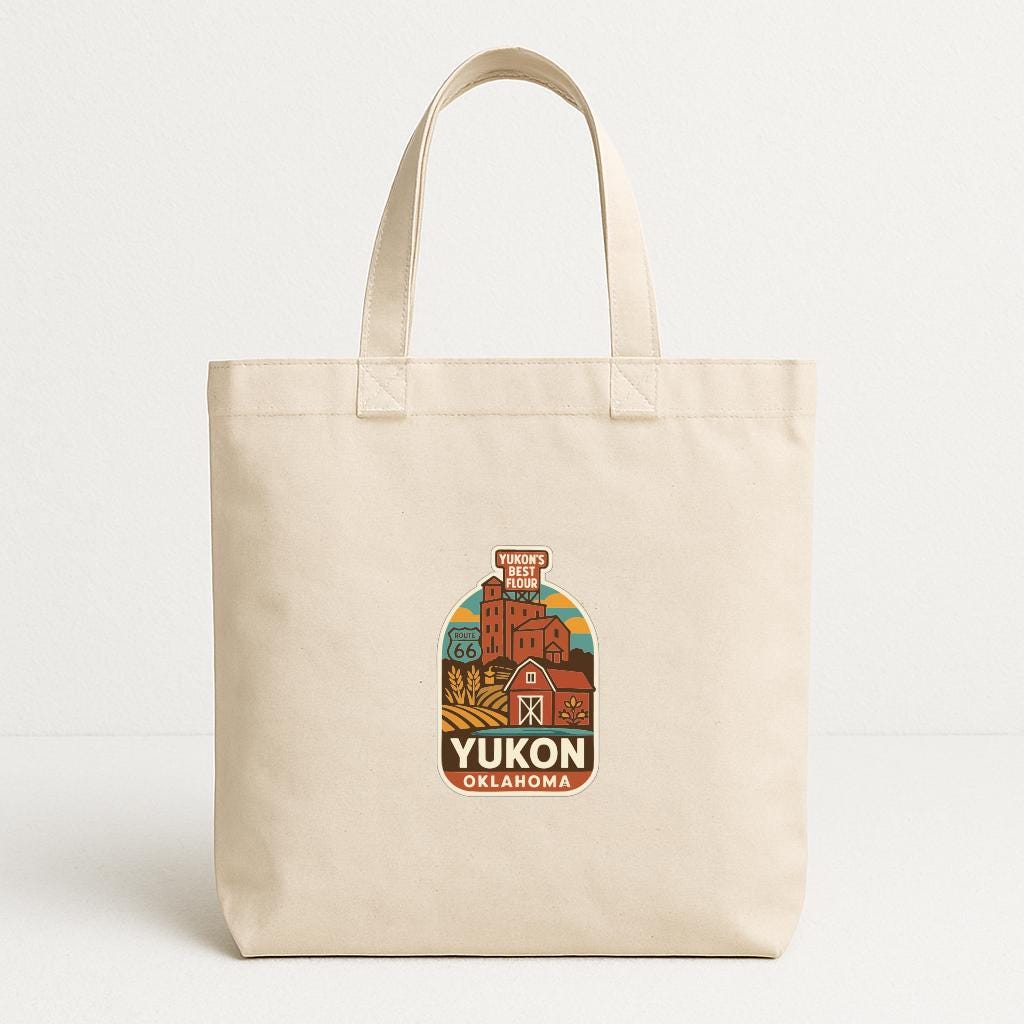 A beige tote bag with a printed design depicting a rural landscape and the text "YUKON OKLAHOMA" printed on it.
