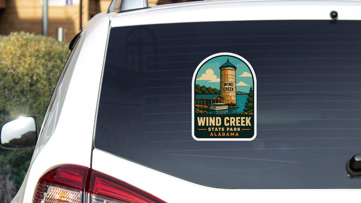 A sticker on the rear window of a white vehicle, depicting a scenic image of Wind Creek State Park in Alabama.