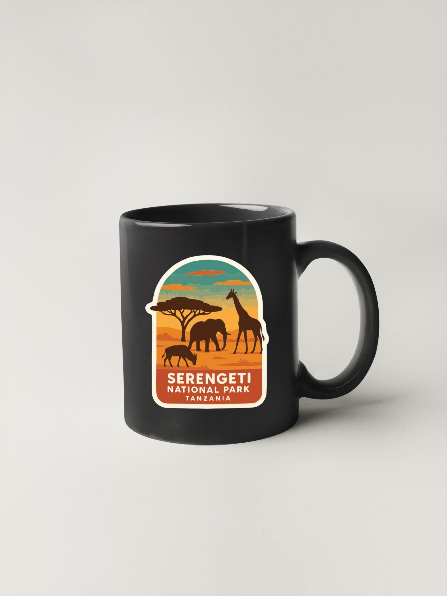 A black coffee mug with a sticker depicting wildlife silhouettes and the text "Serengeti National Park, Tanzania".