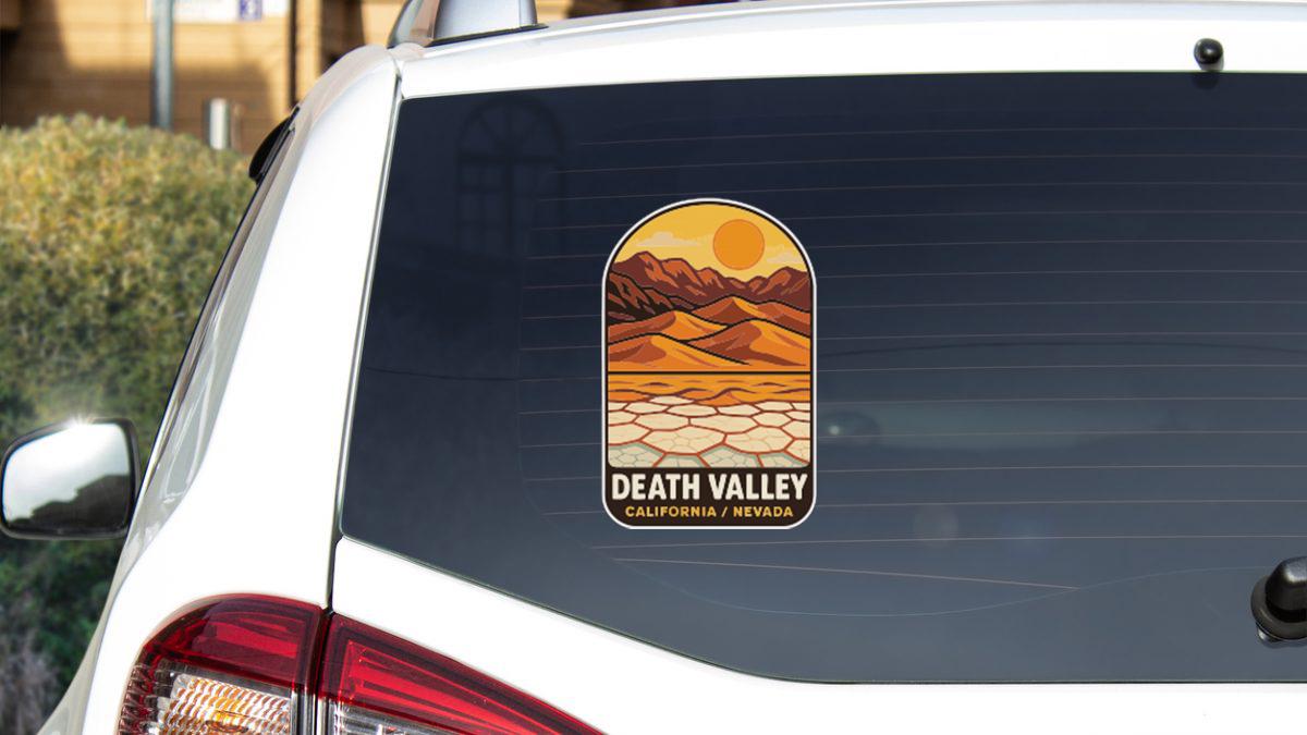 A sticker on the rear window of a white car, depicting a desert landscape with mountains and a sun in the sky.