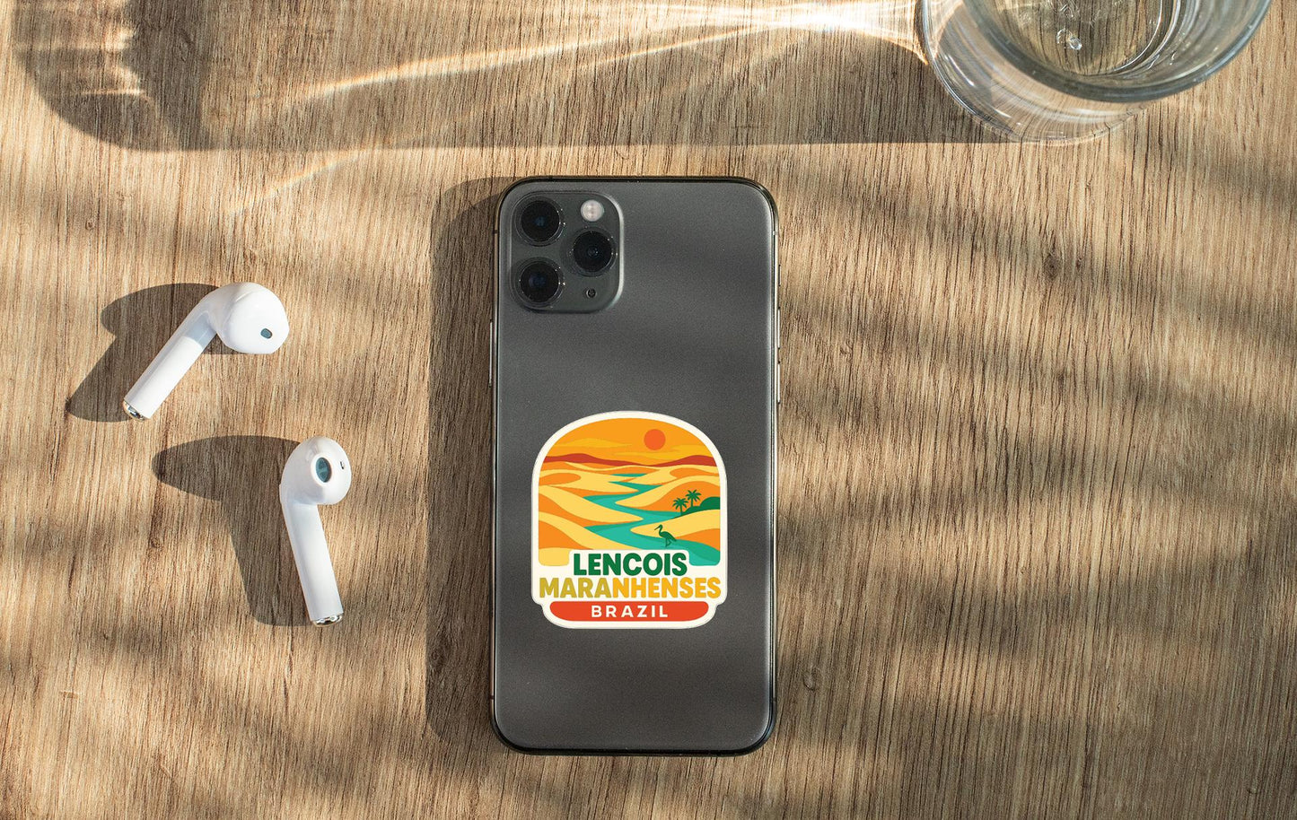 a smartphone with a sticker on the back, placed on a wooden surface alongside a pair of white earbuds and a glass of water.
