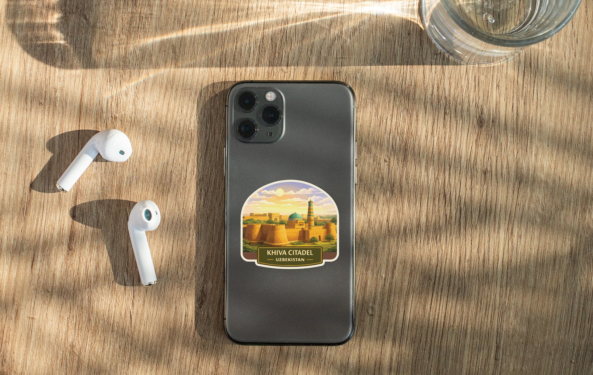 a smartphone with a sticker of a cityscape on its back, along with a pair of white earbuds and a glass of water on a wooden surface.