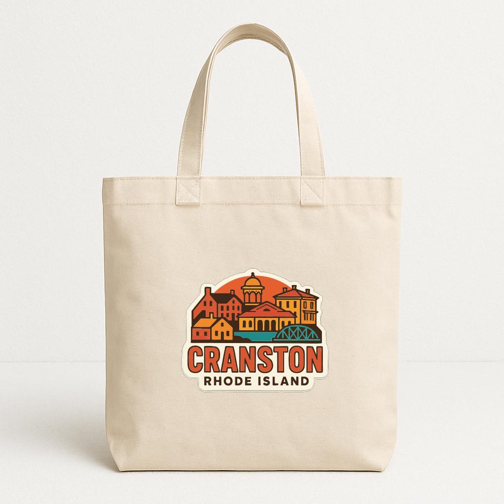 A beige tote bag with a printed design of Cranston, Rhode Island, featuring colorful buildings and a sunset-like background.
