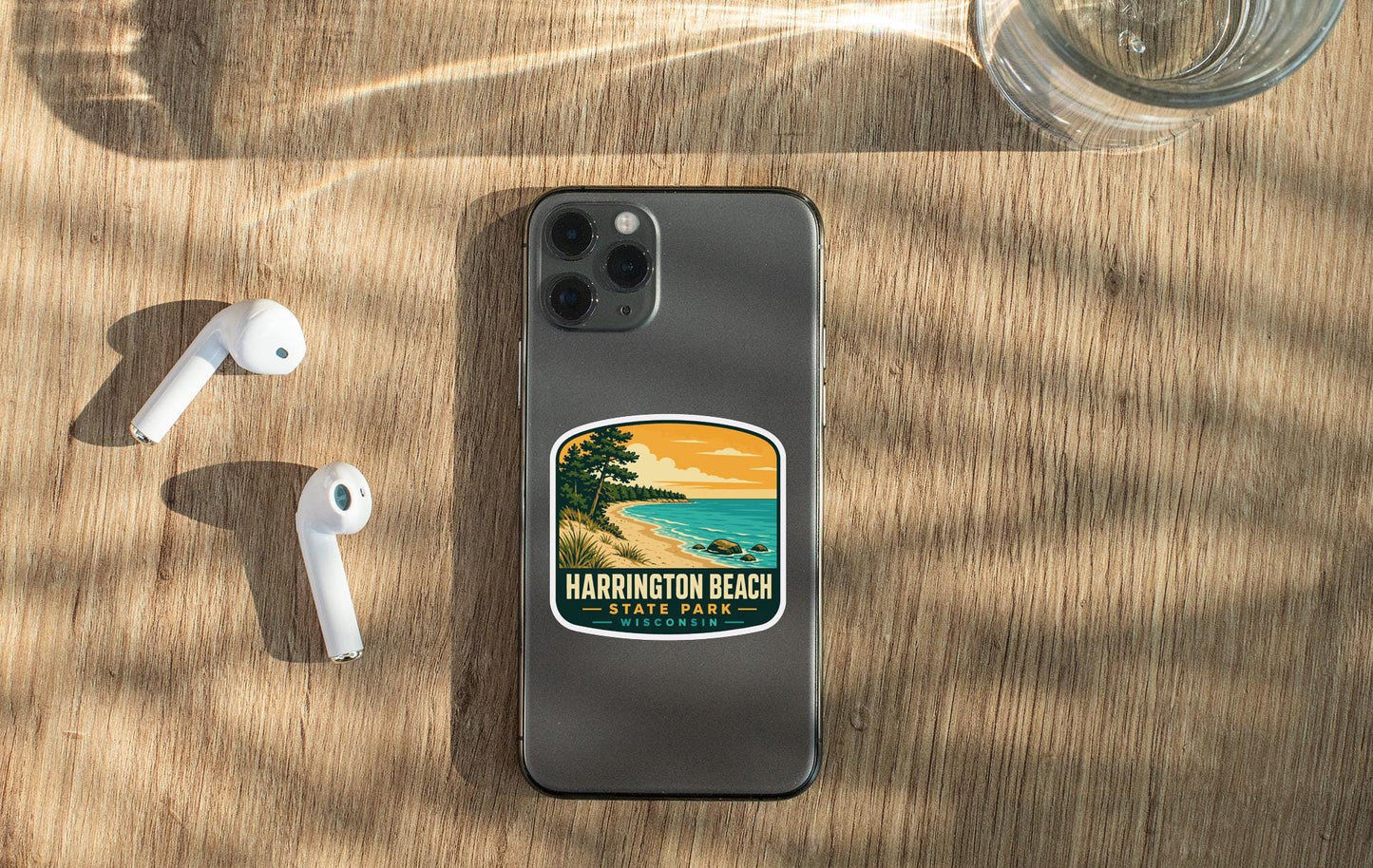 a smartphone with a sticker on the back, placed on a wooden surface alongside a pair of white earbuds and a glass of water.