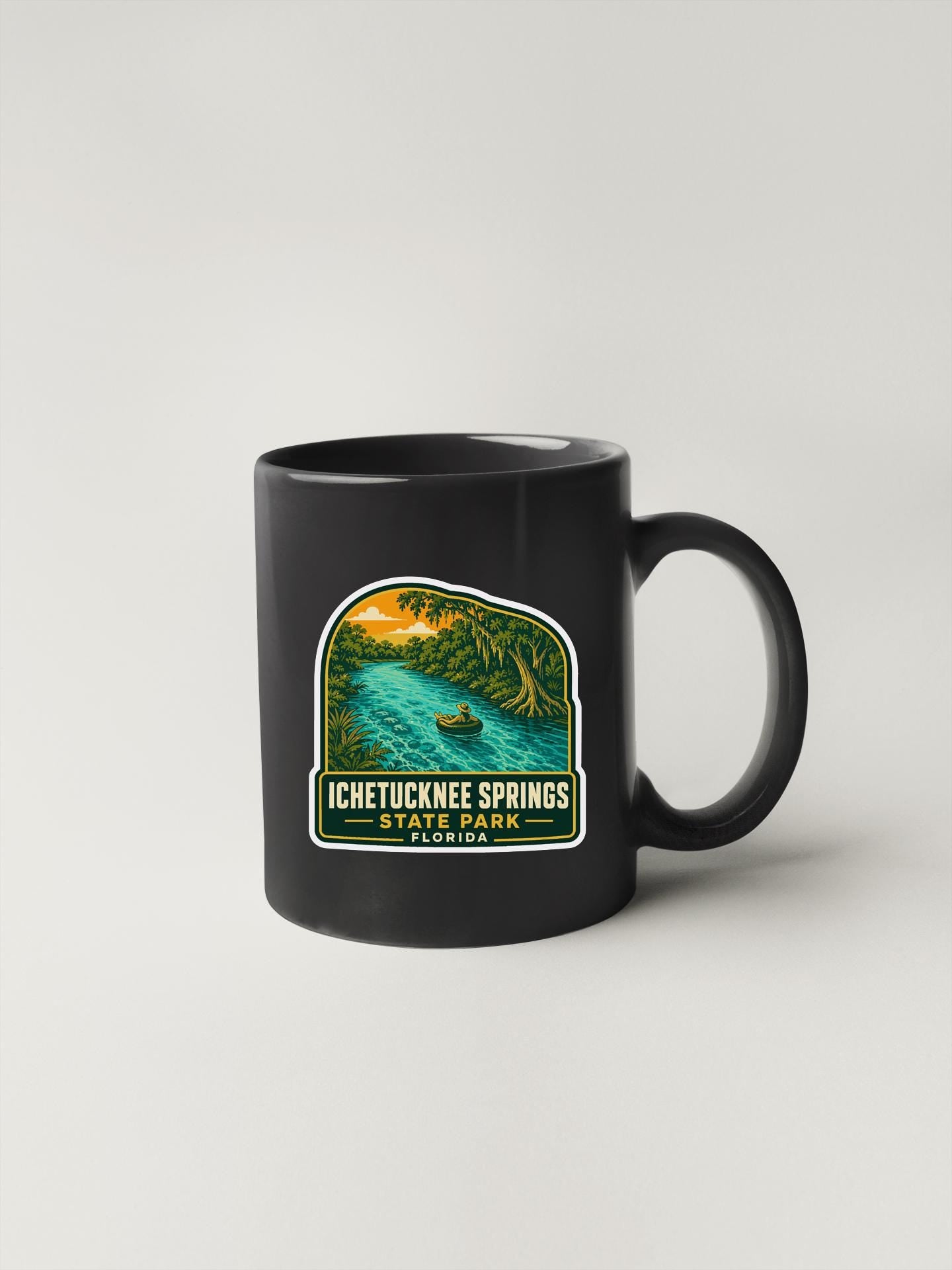 A black coffee mug with a sticker depicting a scenic landscape featuring a river and palm trees.