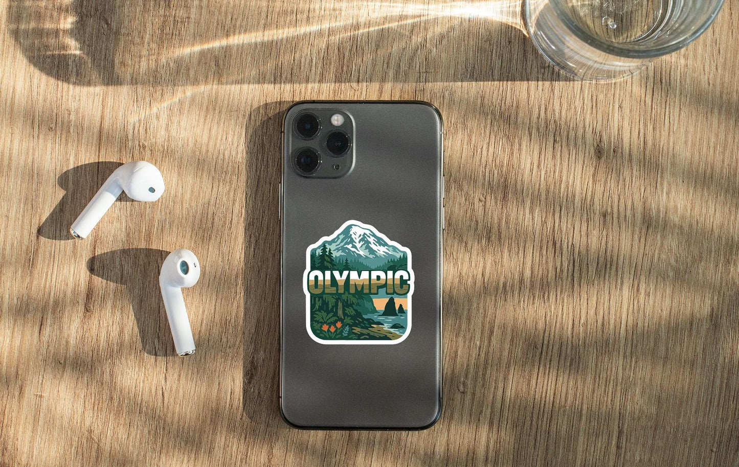 a smartphone with a sticker on the back, placed on a wooden surface alongside a pair of white earbuds and a glass of water.