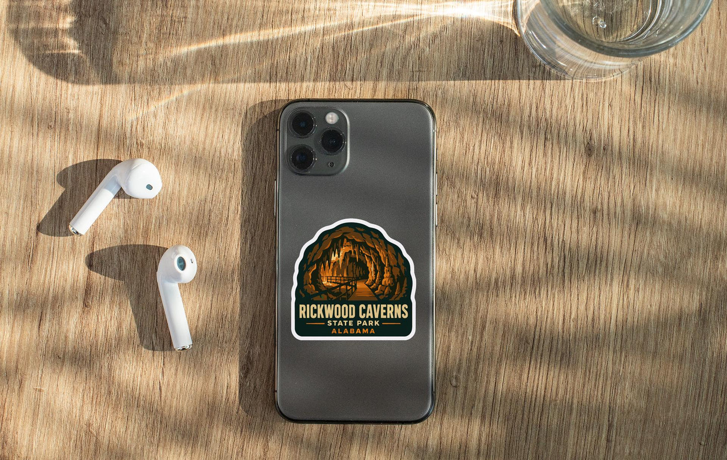 a smartphone with a sticker on it, placed on a wooden surface alongside a pair of white earbuds and a glass of water.