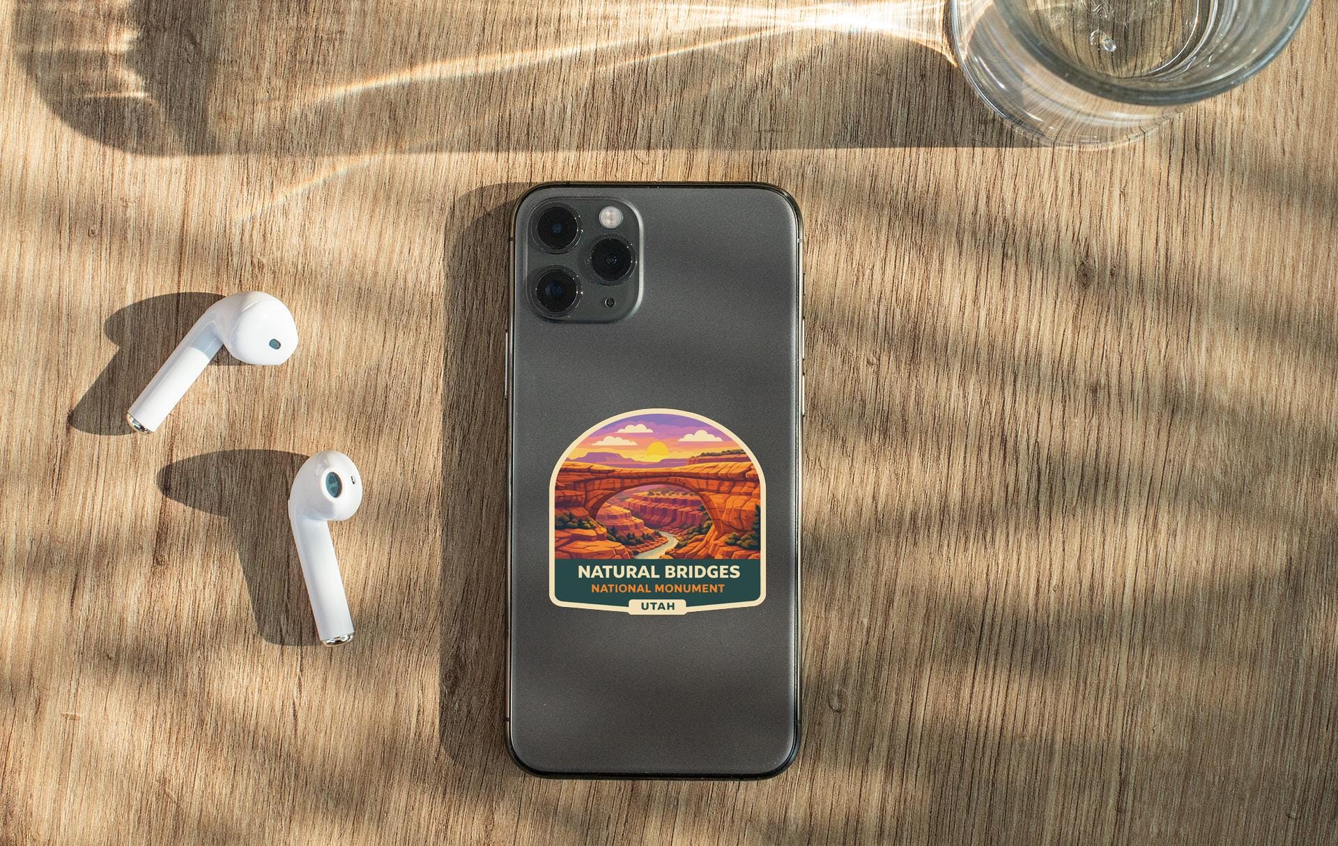 a smartphone with a sticker of a scenic natural bridge, a pair of white earbuds, and a glass of water on a wooden surface.