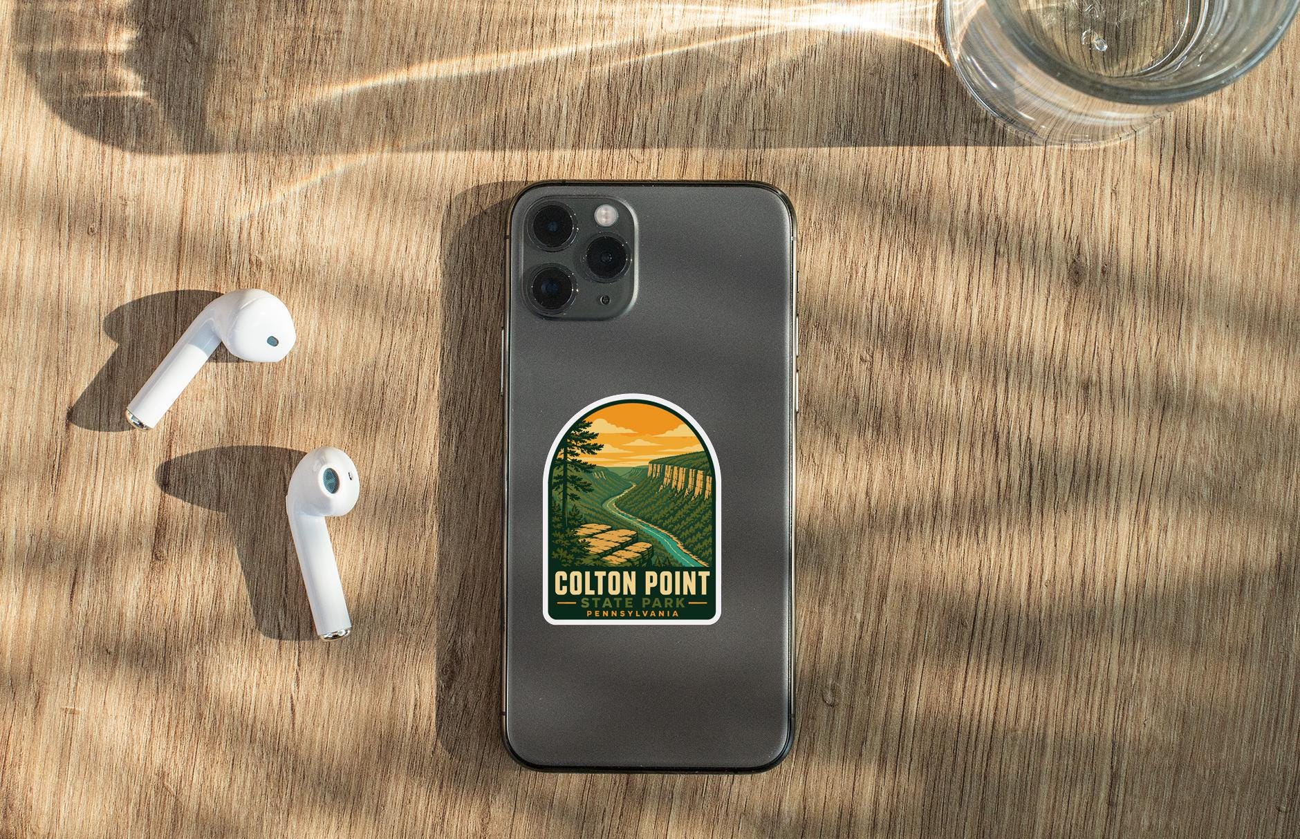 a smartphone with a sticker on it, placed on a wooden surface alongside a pair of white earbuds and a glass of water.