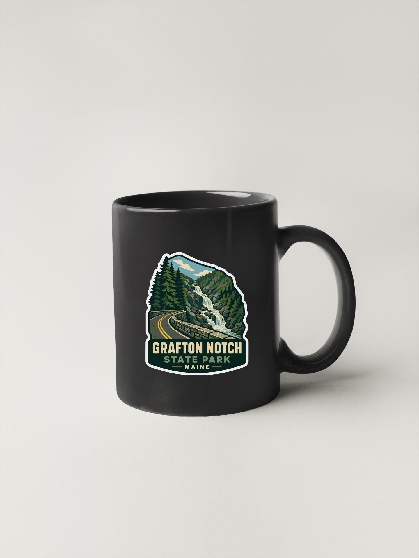 A black coffee mug with a graphic design depicting a scenic mountain landscape, including a waterfall and forest.