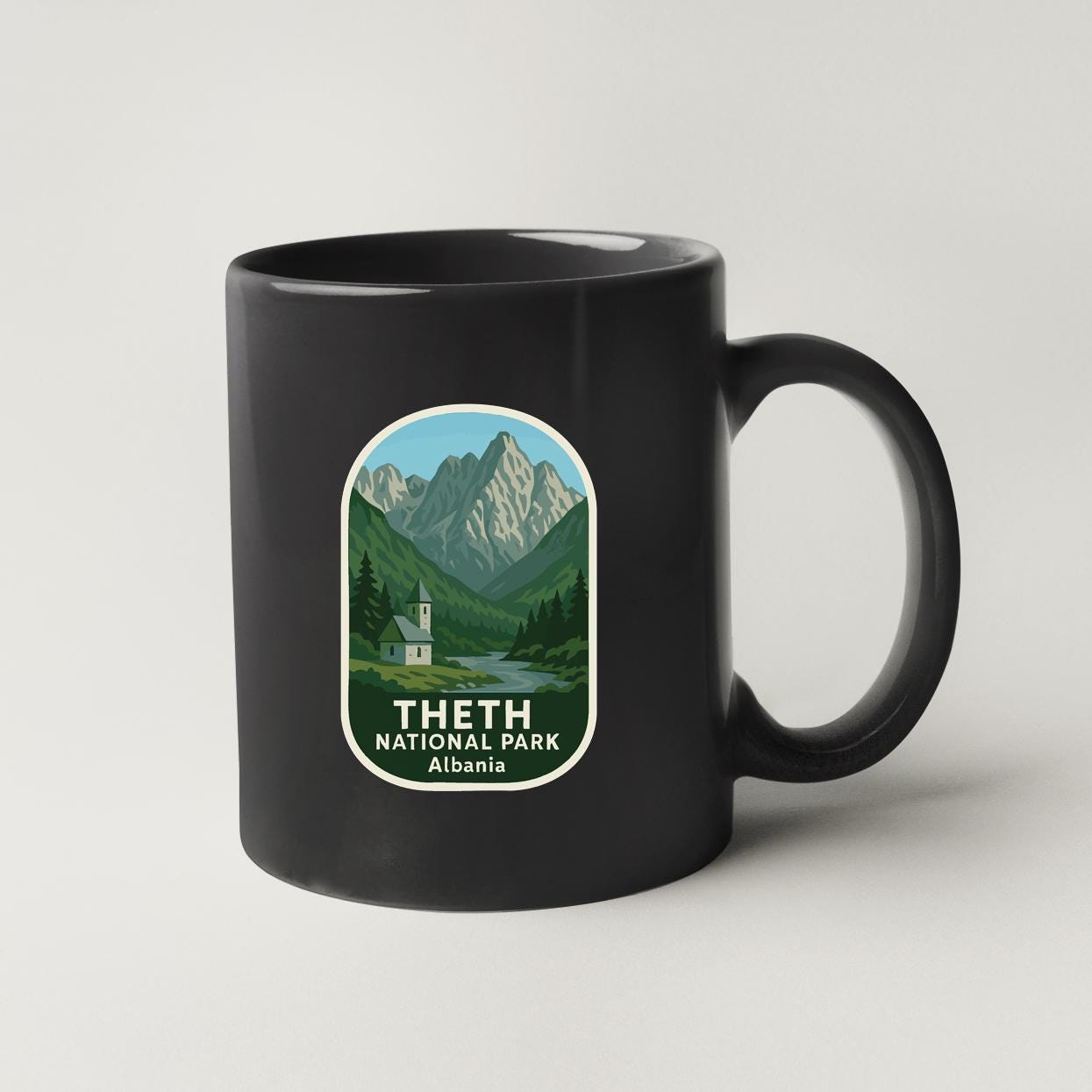 A black coffee mug with a label that features a mountain landscape and the text "THETH NATIONAL PARK ALBANIA".