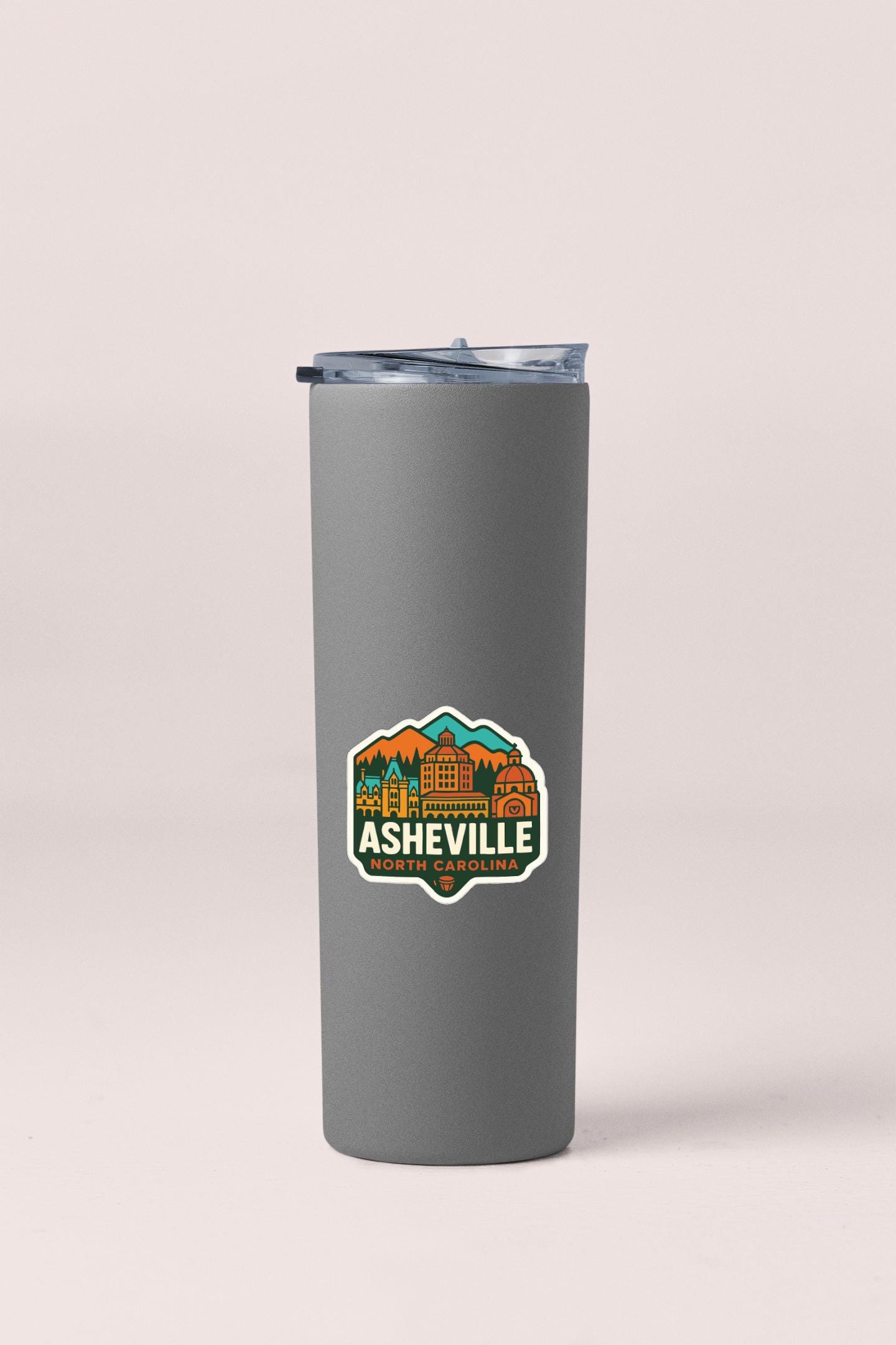 A gray insulated tumbler with a lid and a sticker featuring the text "Ahead of Asheville, NC" and an illustration of a mountain range.