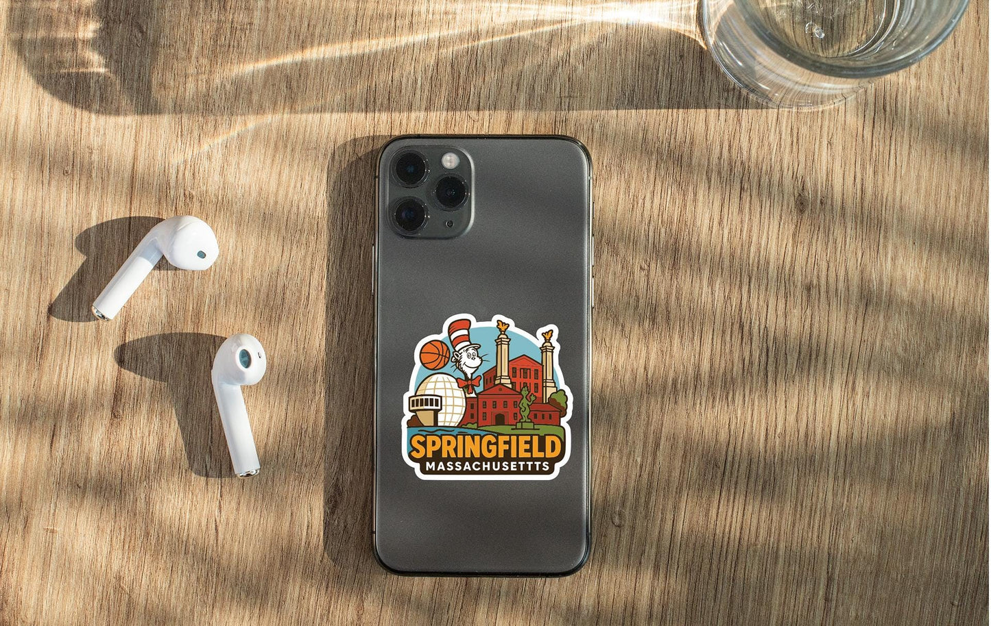 a smartphone with a sticker of the town of Springfield, Massachusetts, placed on a wooden surface alongside a pair of white earbuds and a glass of water.