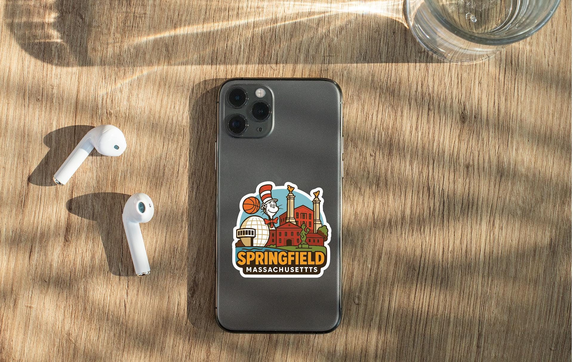 a smartphone with a sticker of the town of Springfield, Massachusetts, placed on a wooden surface alongside a pair of white earbuds and a glass of water.