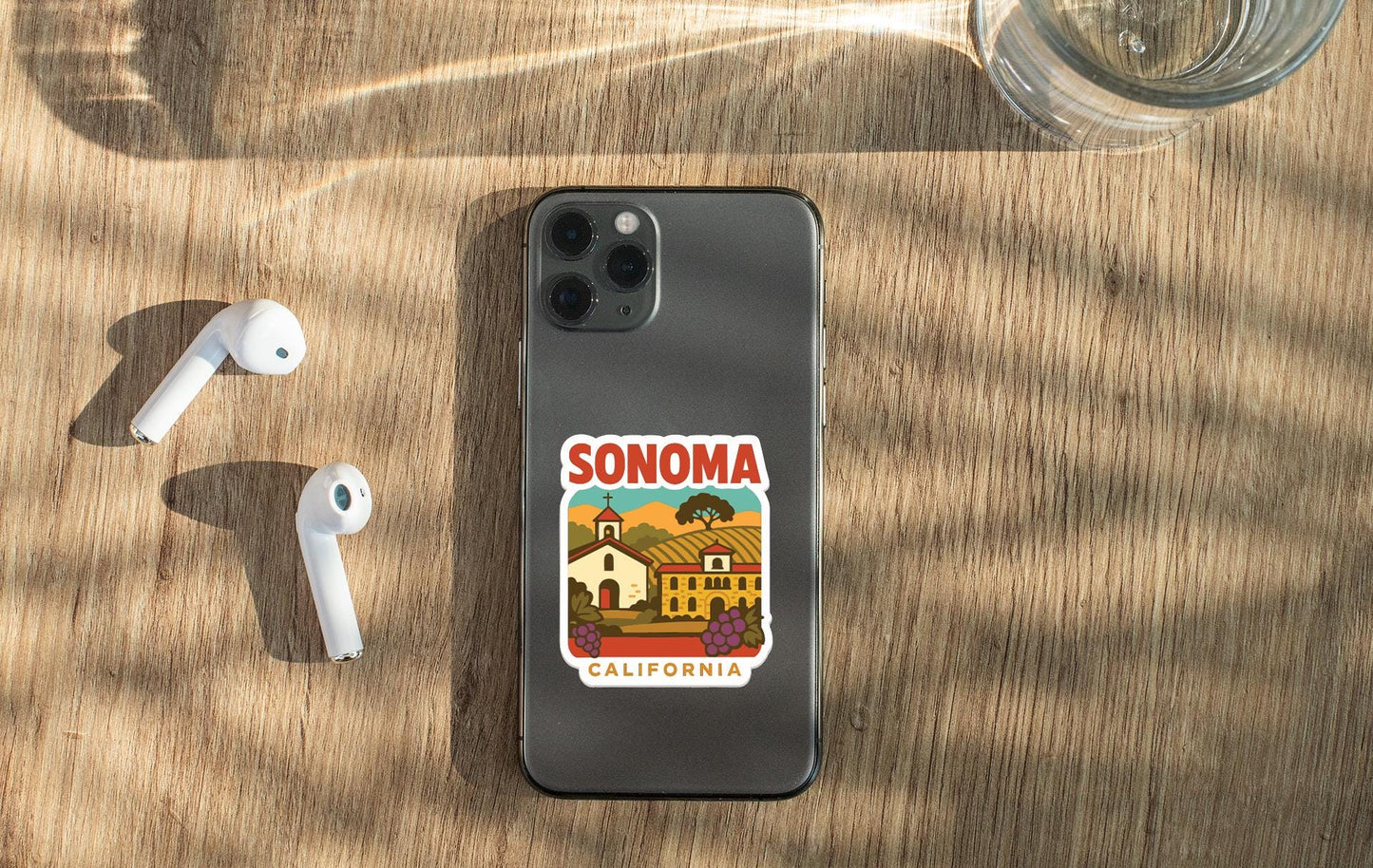 a smartphone with a sticker of Sonoma County, California, placed on a wooden surface alongside a pair of white earbuds and a glass of water.