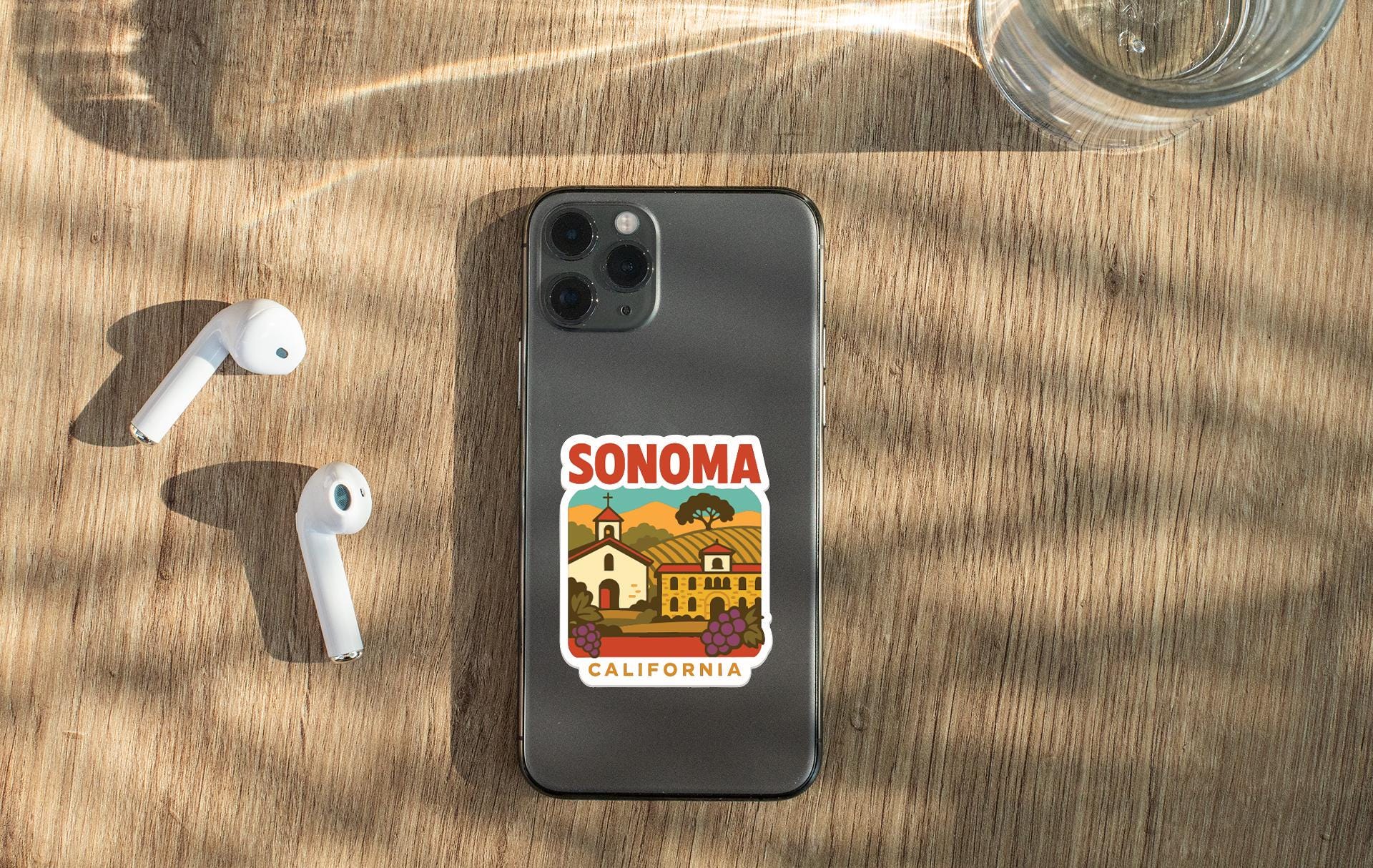 a smartphone with a sticker of Sonoma County, California, placed on a wooden surface alongside a pair of white earbuds and a glass of water.