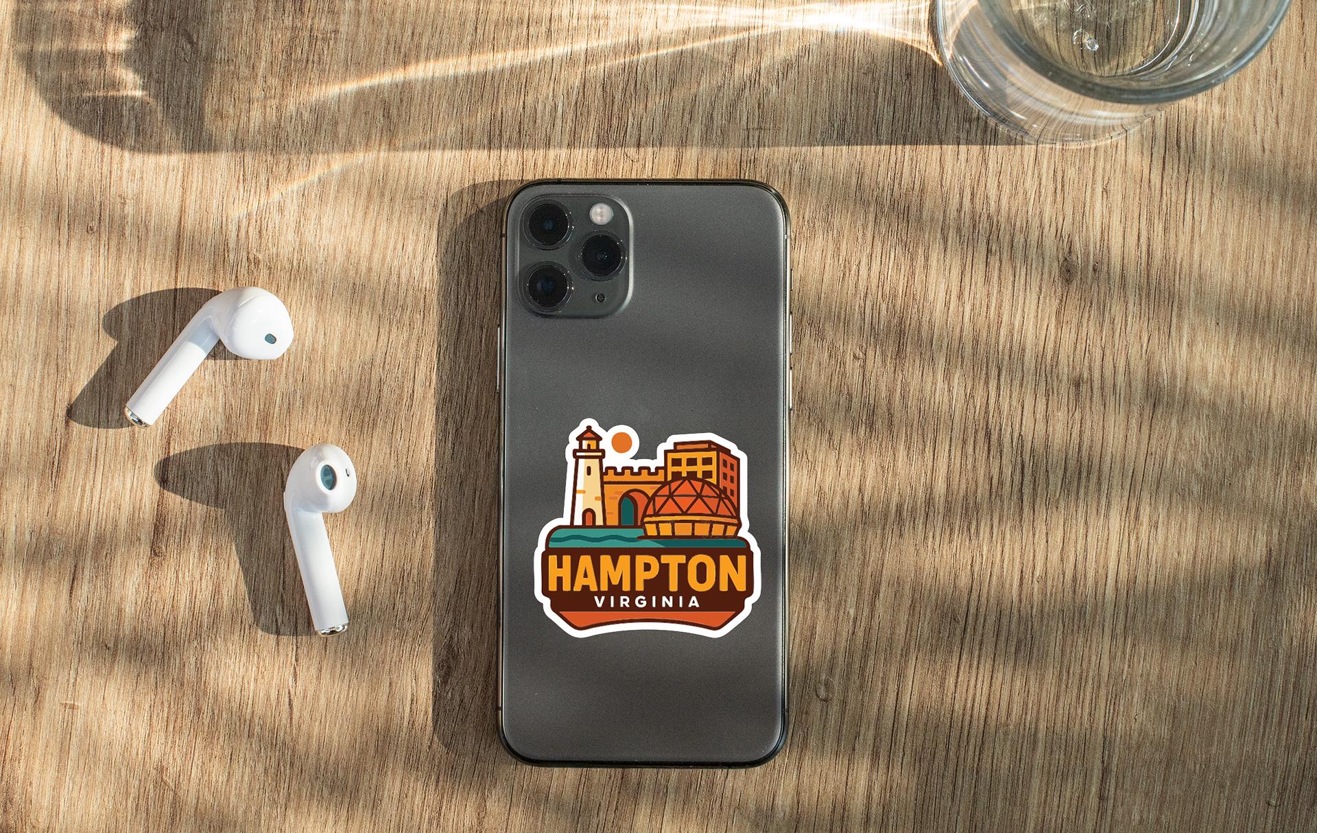 a smartphone with a sticker of Hampton, Virginia, placed on a wooden surface alongside a pair of white earbuds and a glass of water.