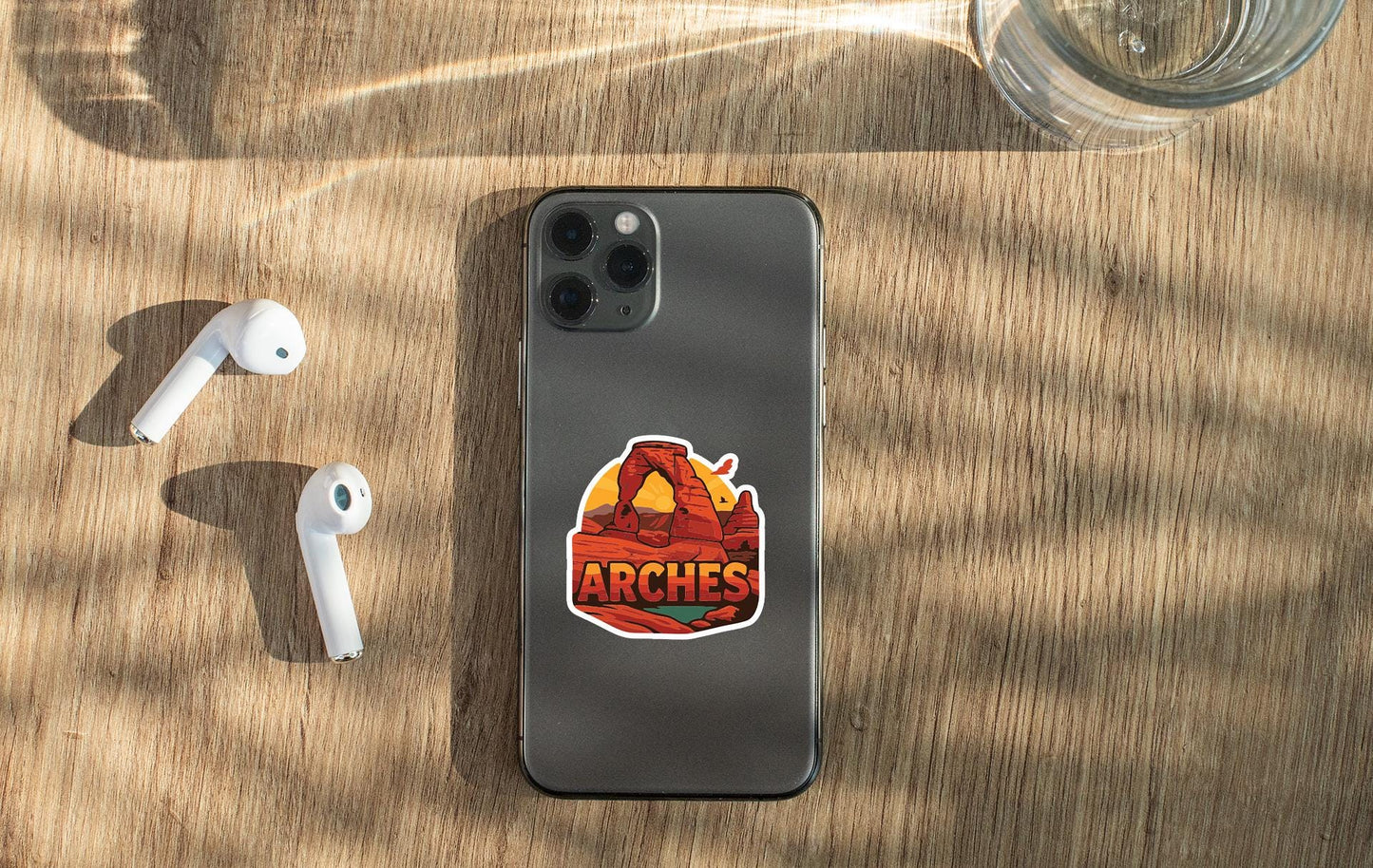 a smartphone with a sticker on the back, placed on a wooden surface alongside a pair of white earbuds and a glass of water.