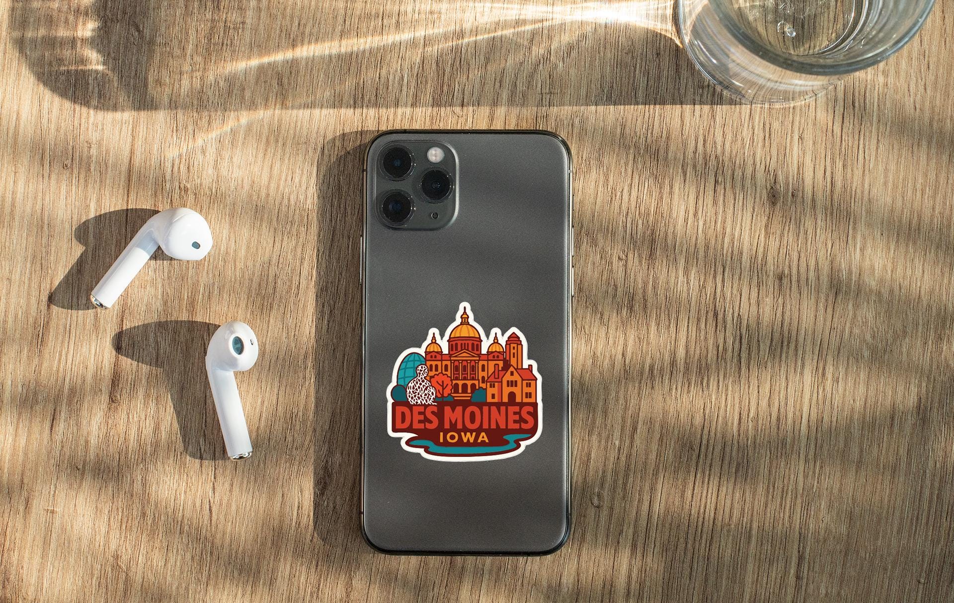 a smartphone with a sticker on the back, placed on a wooden surface alongside a pair of white earbuds and a glass of water.