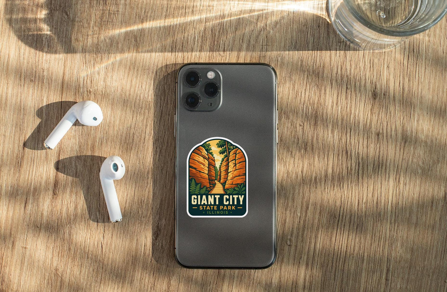 a smartphone with a sticker on the back, placed on a wooden surface alongside a pair of white earbuds and a glass of water.