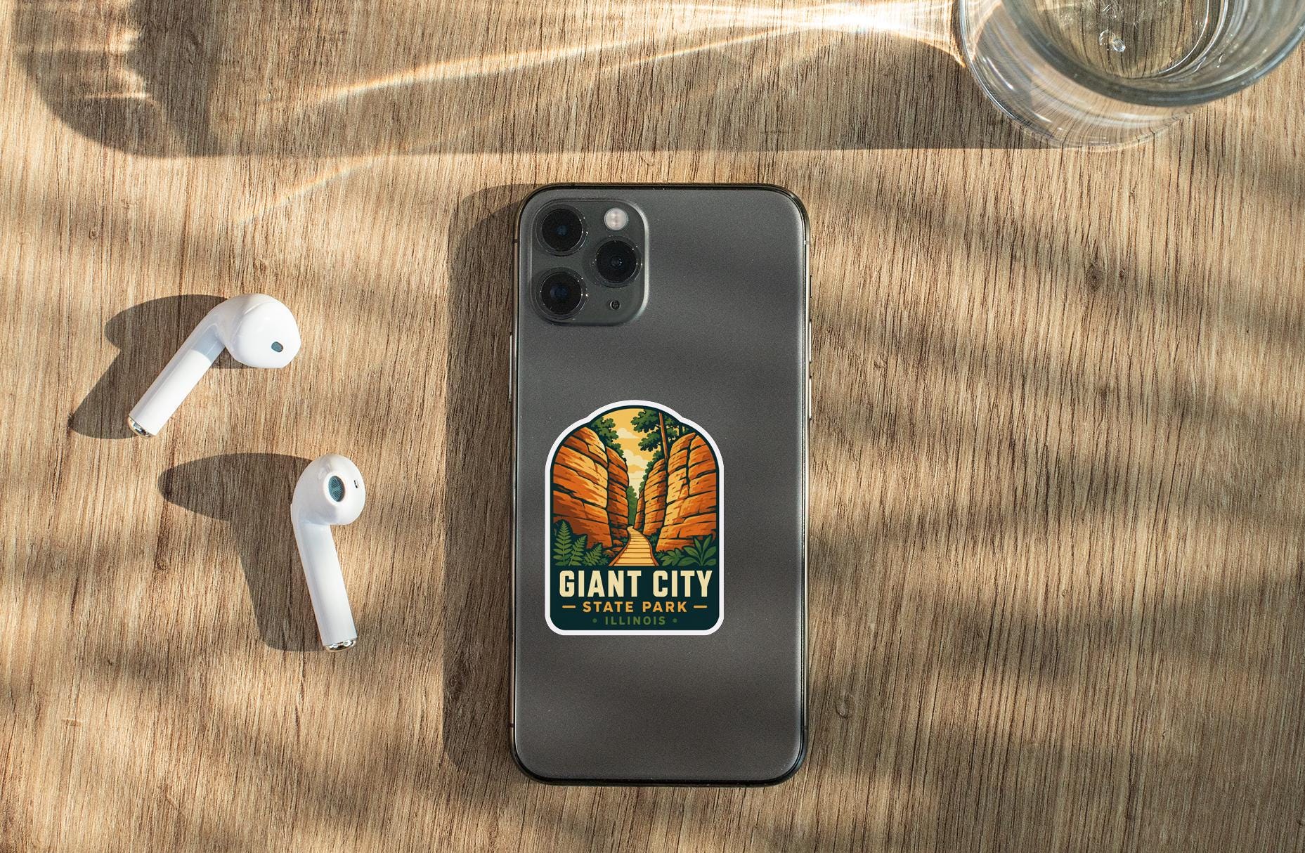 a smartphone with a sticker on the back, placed on a wooden surface alongside a pair of white earbuds and a glass of water.