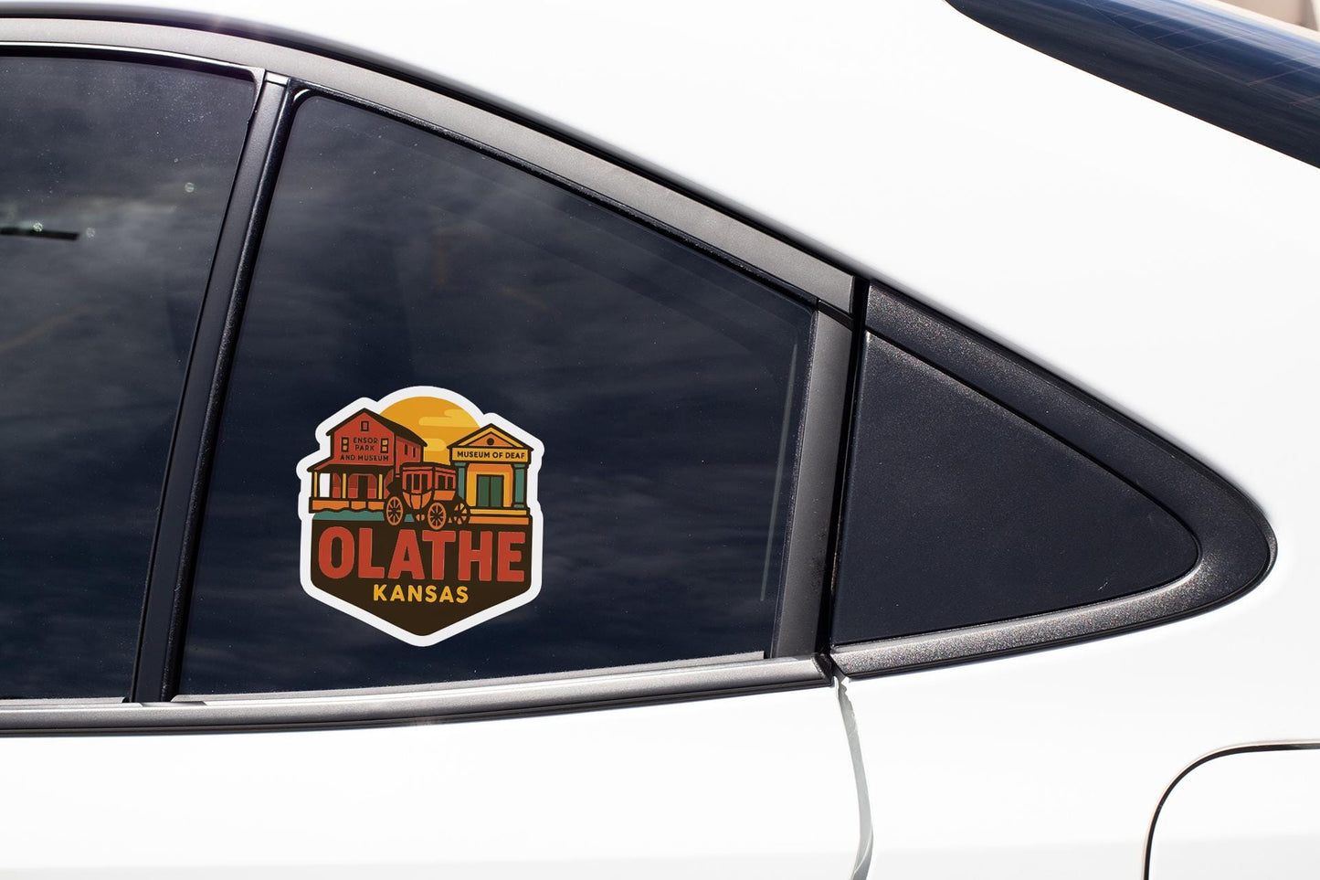 A sticker on the side of a car window, depicting a house with a barn and a sun in the sky, along with the text "OLATHE KANSAS".