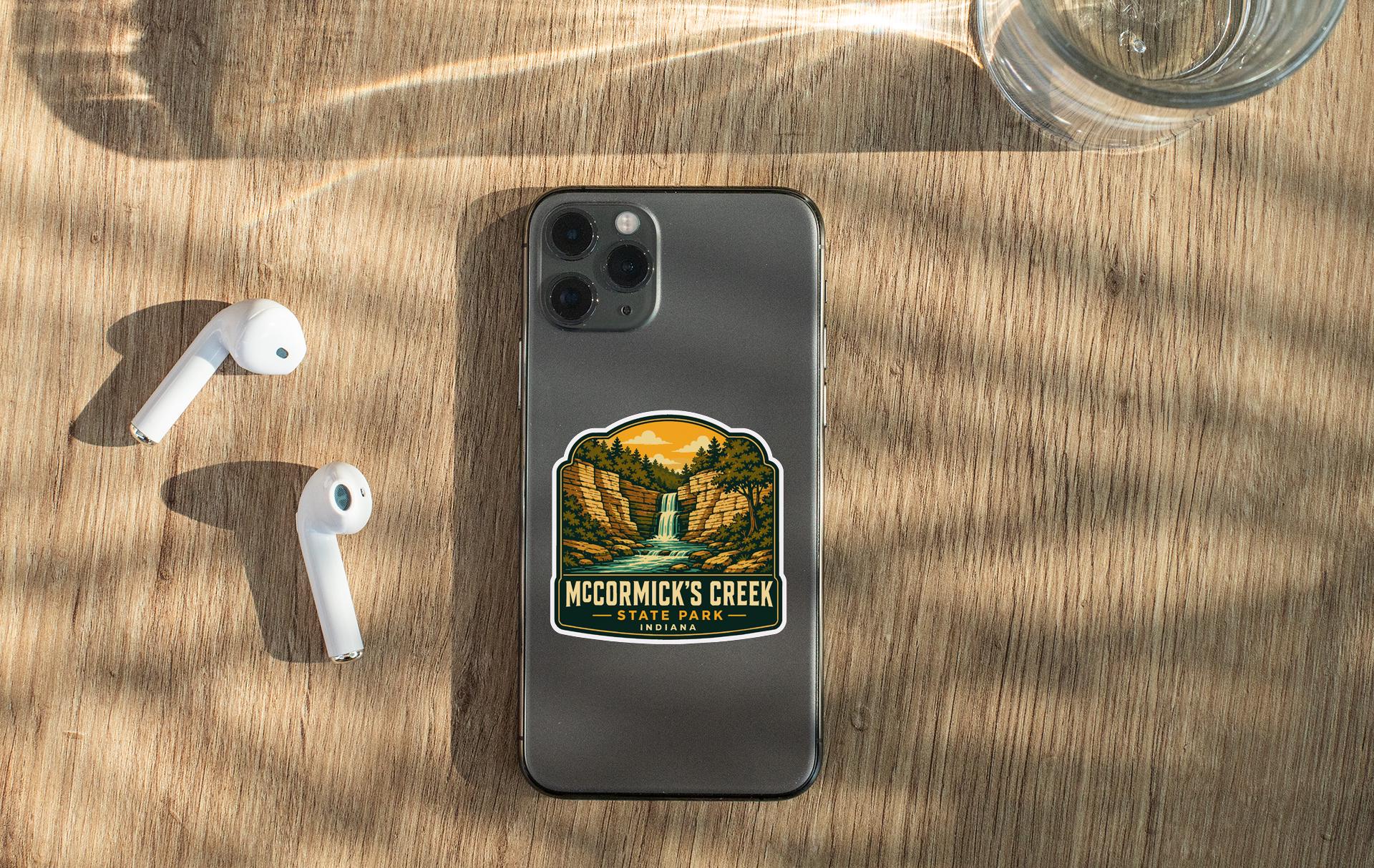 a smartphone with a sticker on the back, placed on a wooden surface alongside a pair of white earbuds and a glass of water.