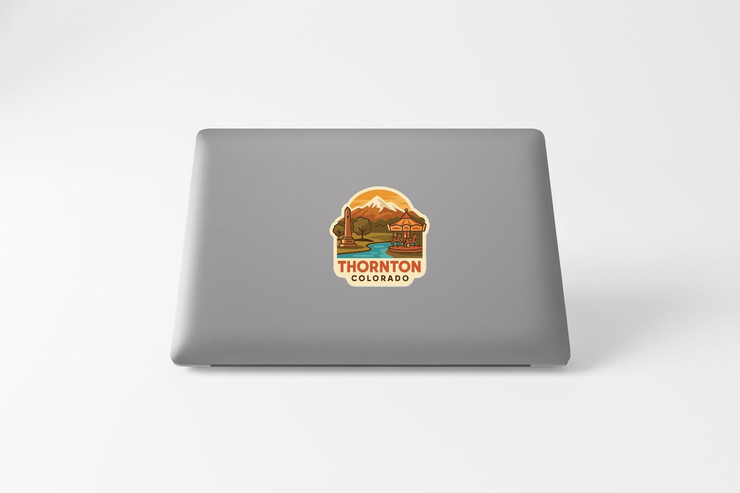 A sticker on the back of a laptop displaying a scenic mountain landscape with a river and a sign that says "THORNTON COLORADO".