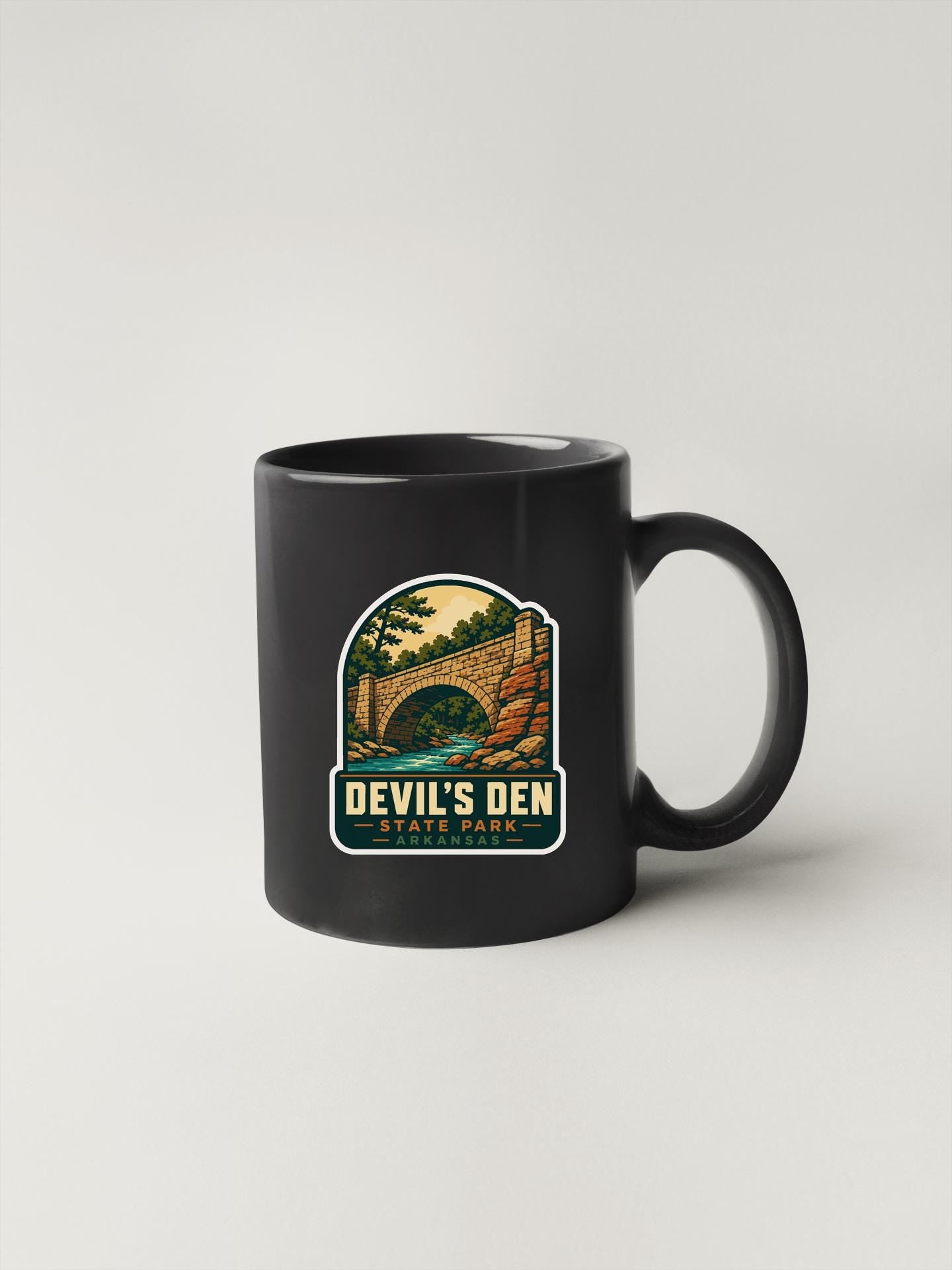 A black coffee mug with a graphic design depicting a bridge and trees, along with the text "Devil's Den State Park" printed on it.