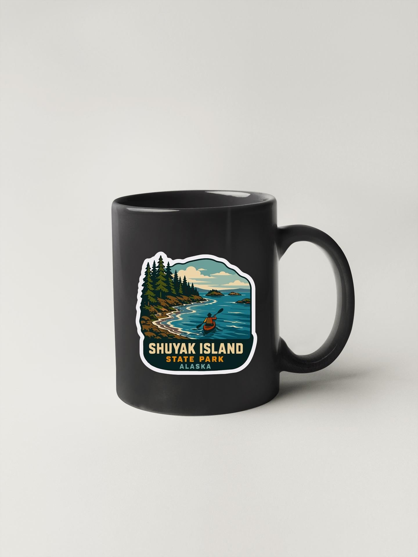 A black coffee mug with a sticker depicting a scenic landscape of Shuyak Island, State Park, Alaska.
