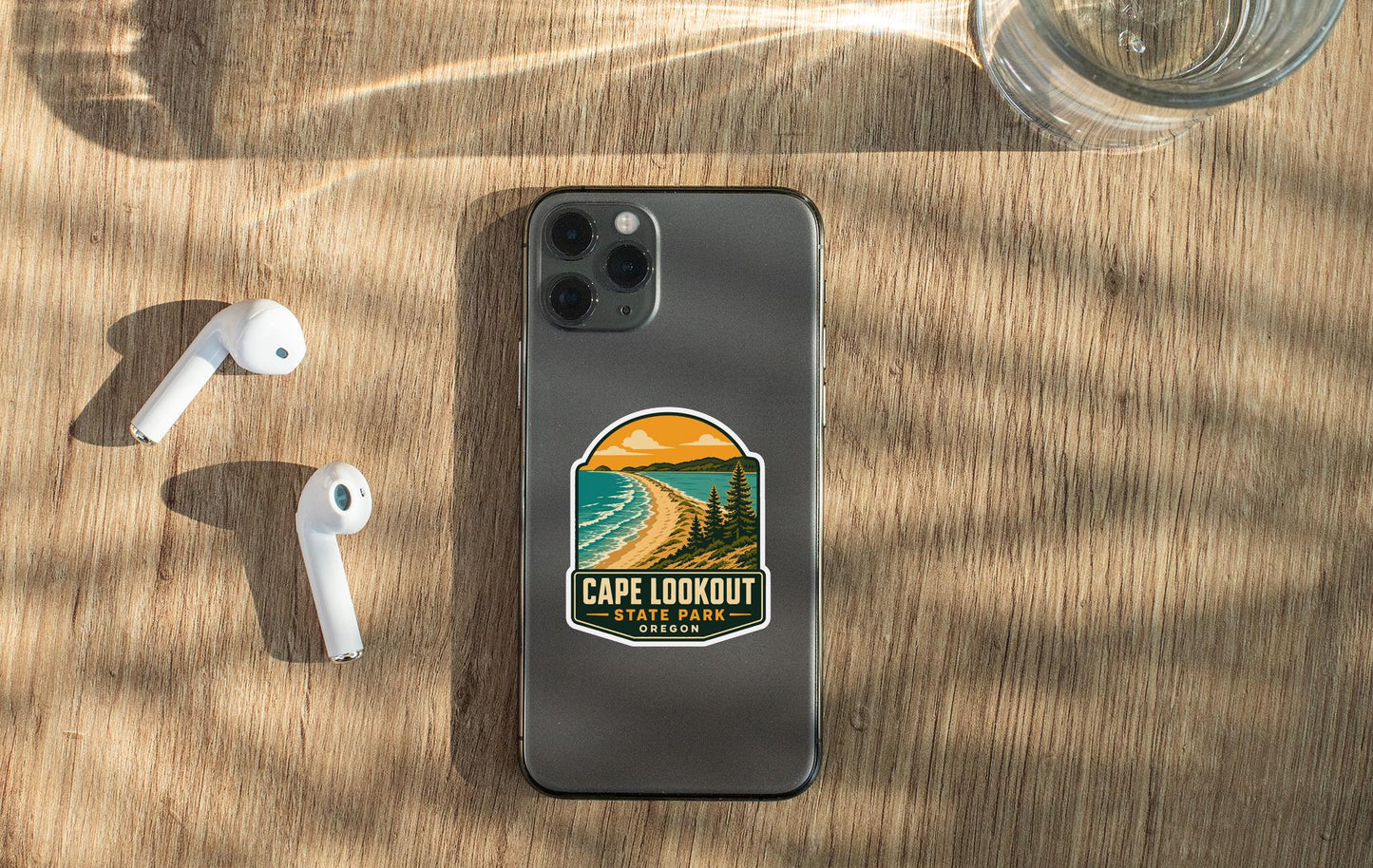 a smartphone with a sticker on the back, placed on a wooden surface alongside a pair of white earbuds and a glass of water.