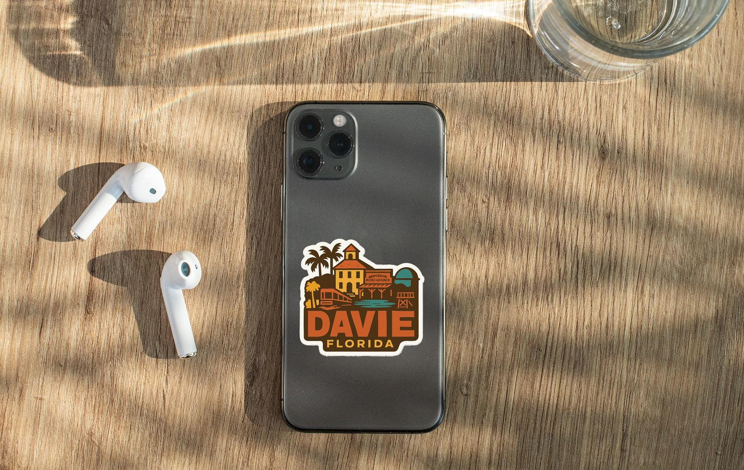 a smartphone with a sticker on the back, placed on a wooden surface alongside a pair of white earbuds and a glass of water.