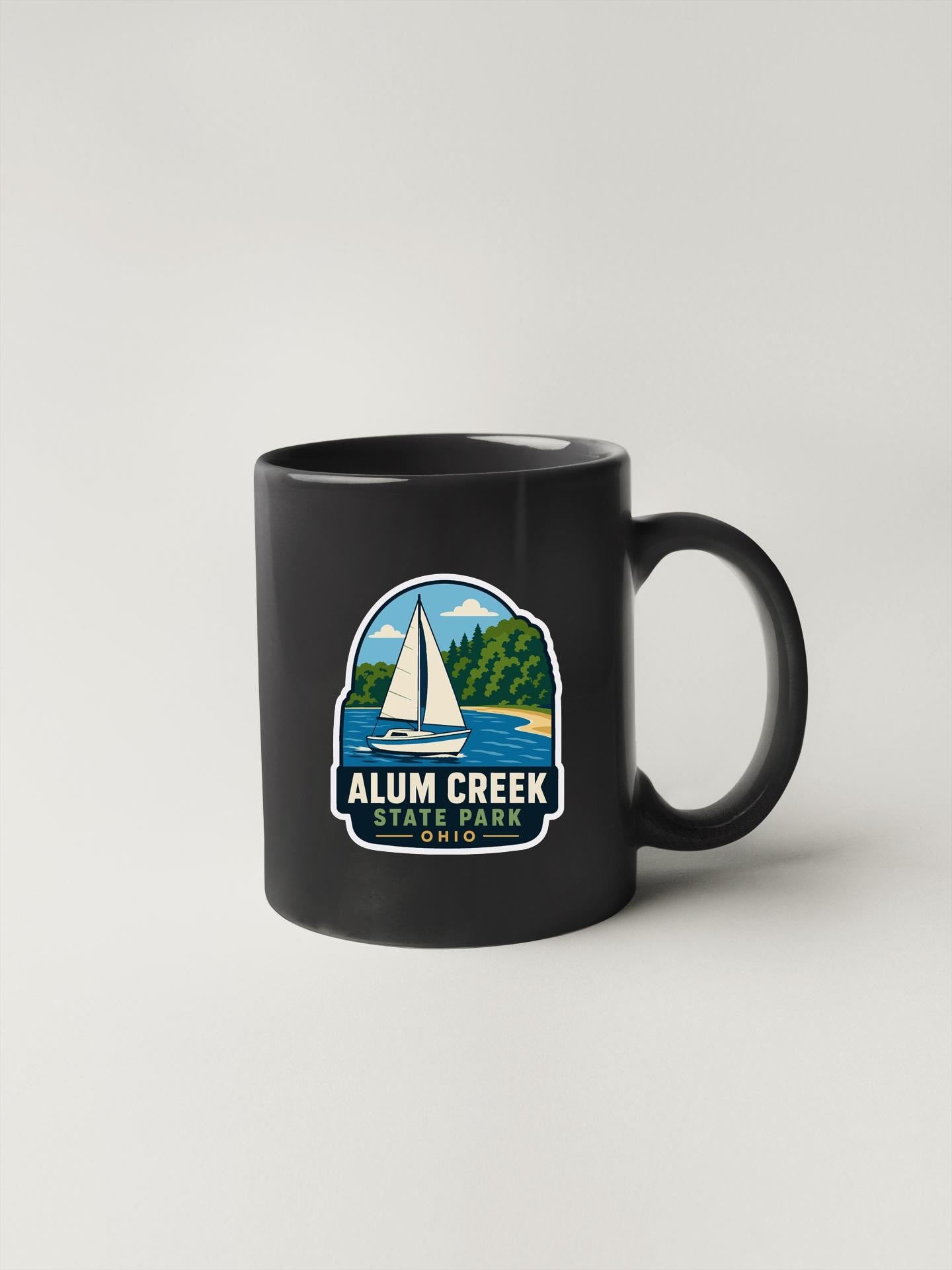 A black coffee mug with a graphic design depicting a sailboat on a lake, surrounded by trees and mountains, with the text "ALUM CREEK STATE PARK" printed on it.