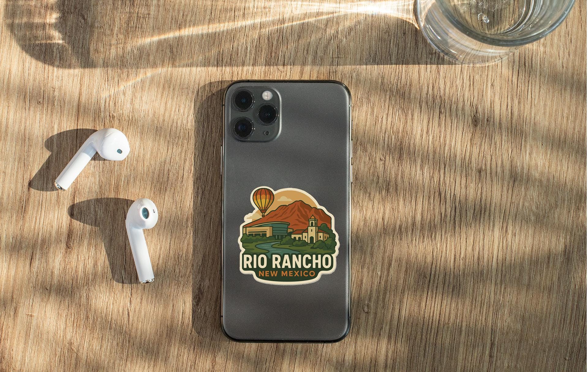 a smartphone with a sticker of Rio Rancho, New Mexico, on its back, along with a pair of white earbuds and a glass of water on a wooden surface.