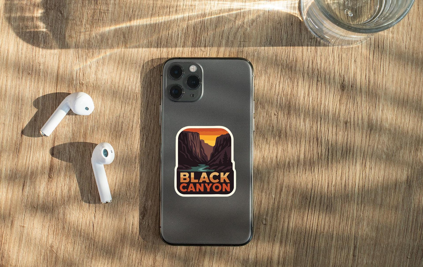 a smartphone with a sticker of the Black Canyon on its back, along with a pair of white earbuds and a glass of water on a wooden surface.