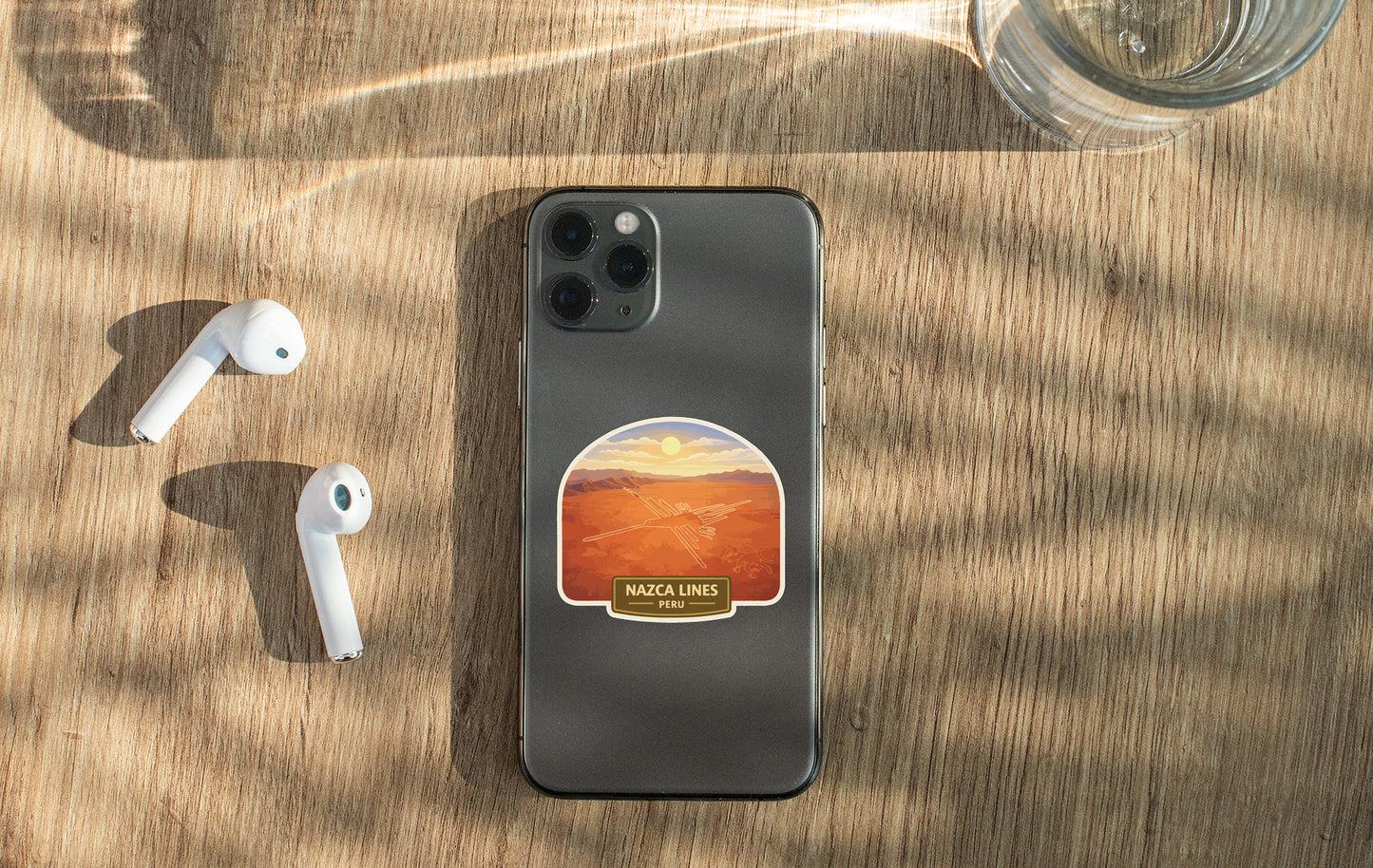a smartphone with a sticker on the back, along with a pair of white earbuds and a glass of water on a wooden surface.