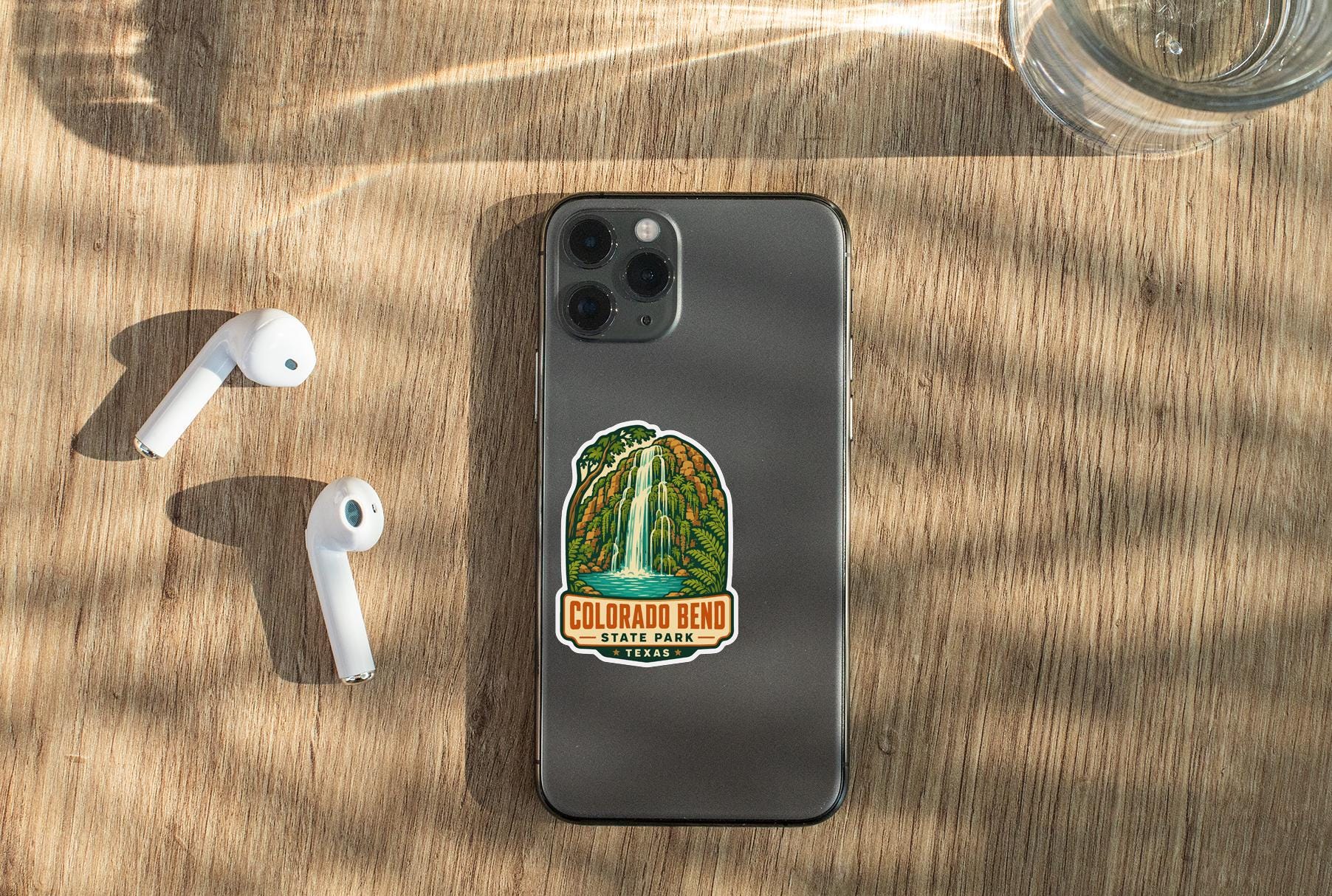 a smartphone with a sticker on it, placed on a wooden surface alongside a pair of white earbuds and a glass of water.
