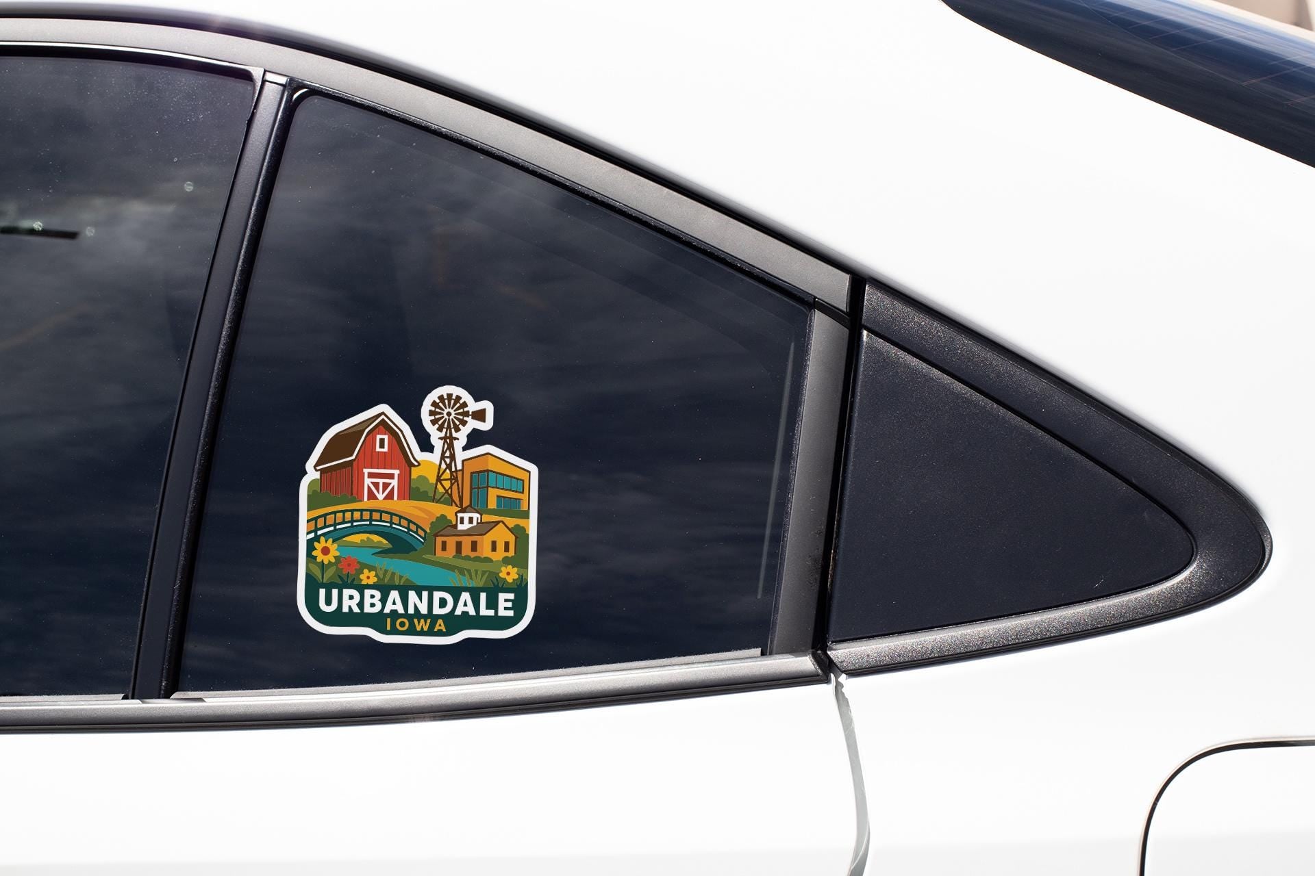 A sticker on the side of a car window, depicting a scenic landscape with a windmill and a barn, representing the town of Urbandale, Iowa.