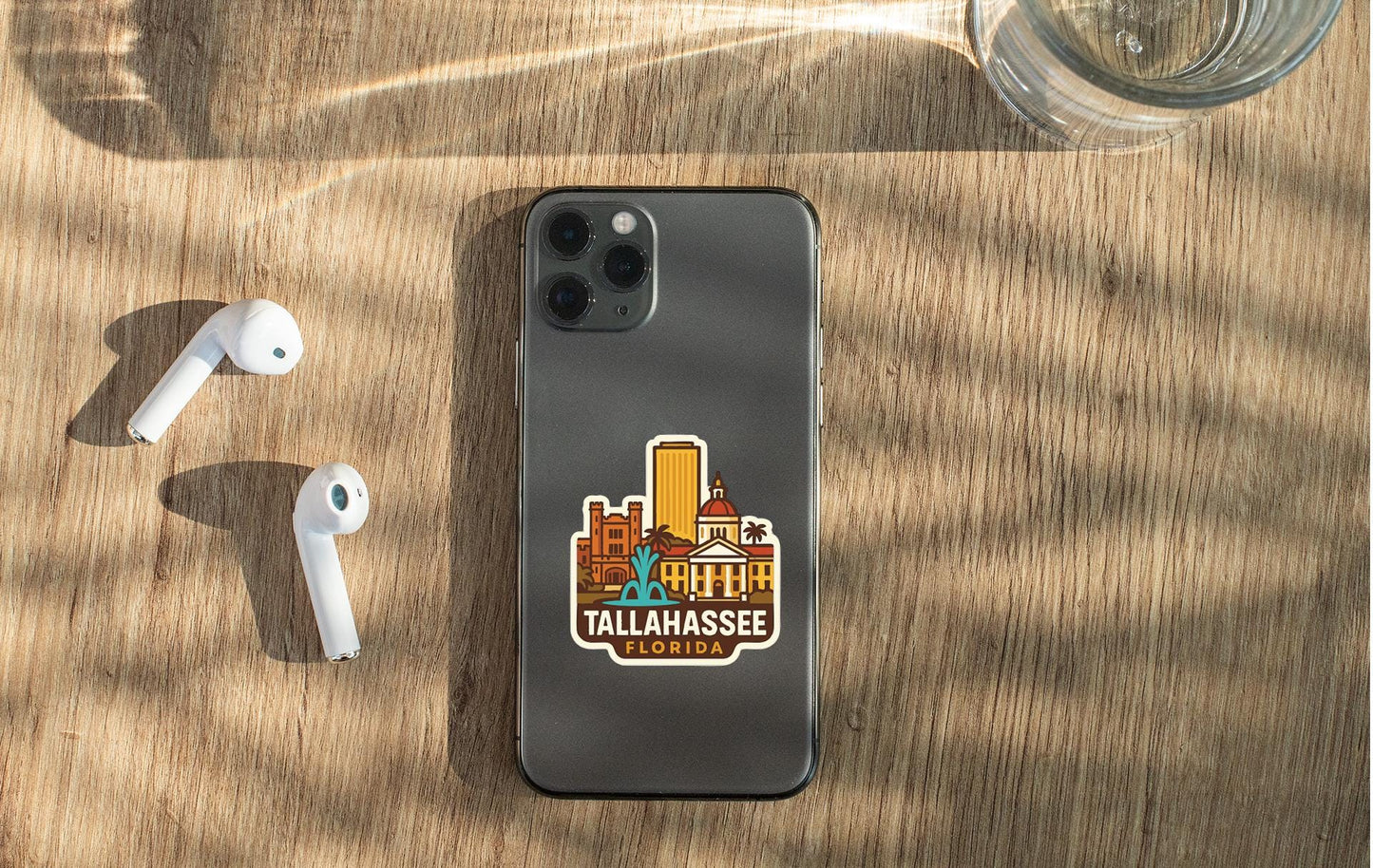 a smartphone with a sticker of Tallahassee, Florida, placed on a wooden surface alongside a pair of white earbuds and a glass of water.