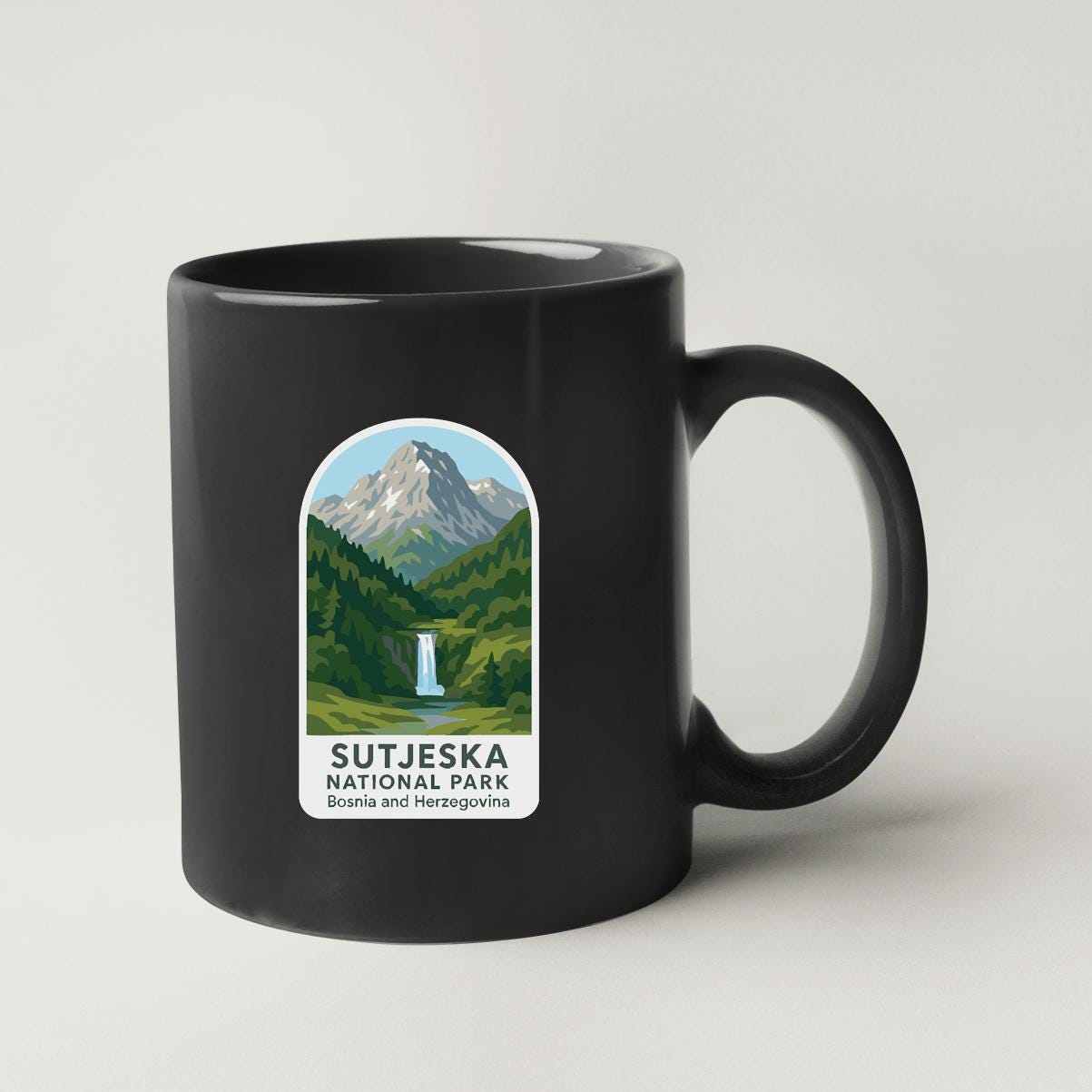 A black coffee mug with a graphic design depicting a mountain landscape and a waterfall, along with the text "SUTJESKA NATIONAL PARK" printed on it.