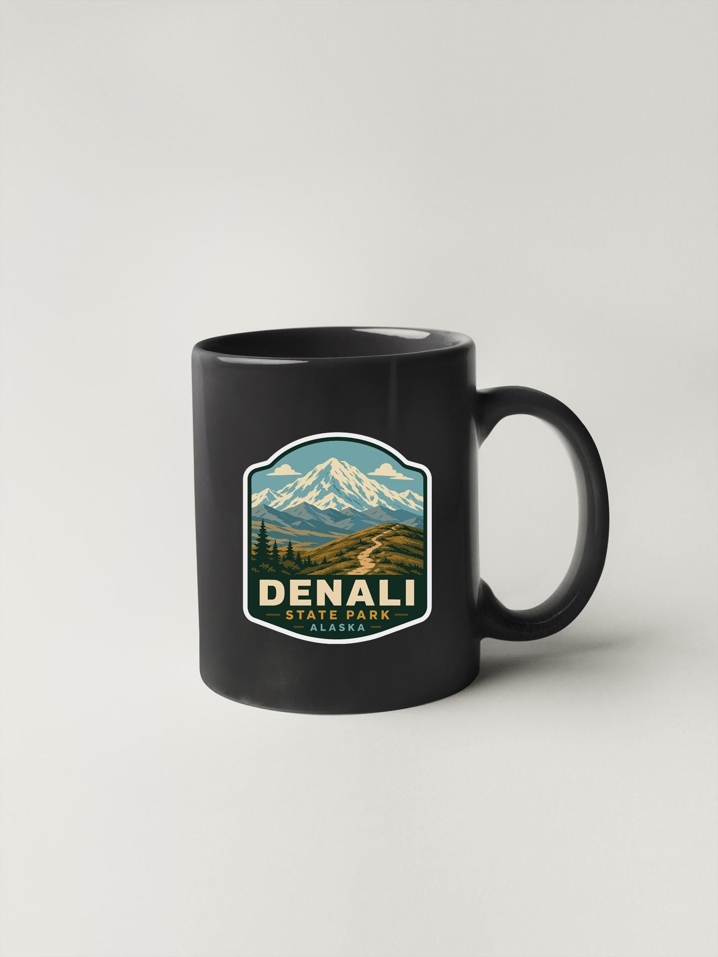 A black coffee mug with a mountain landscape sticker on it, featuring the text "DENALI STATE PARK" and a mountain range.