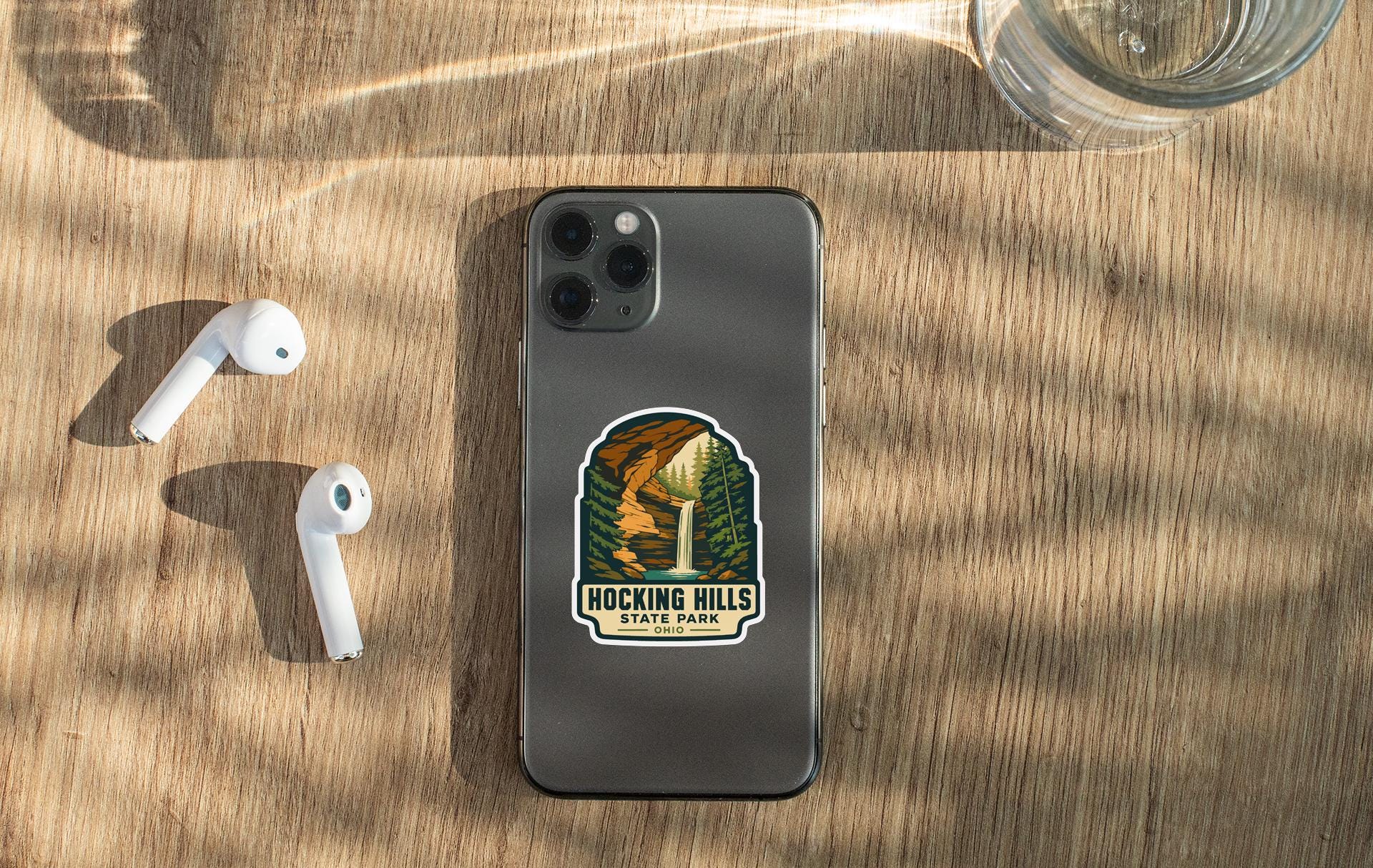 a smartphone with a sticker on it, placed on a wooden surface alongside a pair of white earbuds and a glass of water.
