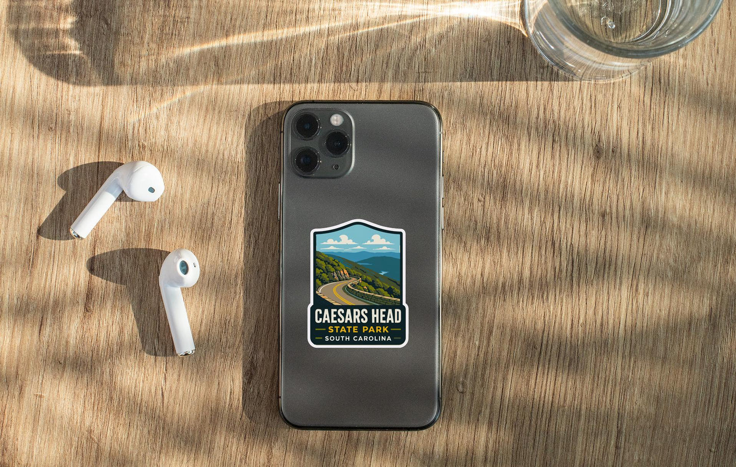 a smartphone with a sticker on the back, placed on a wooden surface alongside a pair of white earbuds and a glass of water.